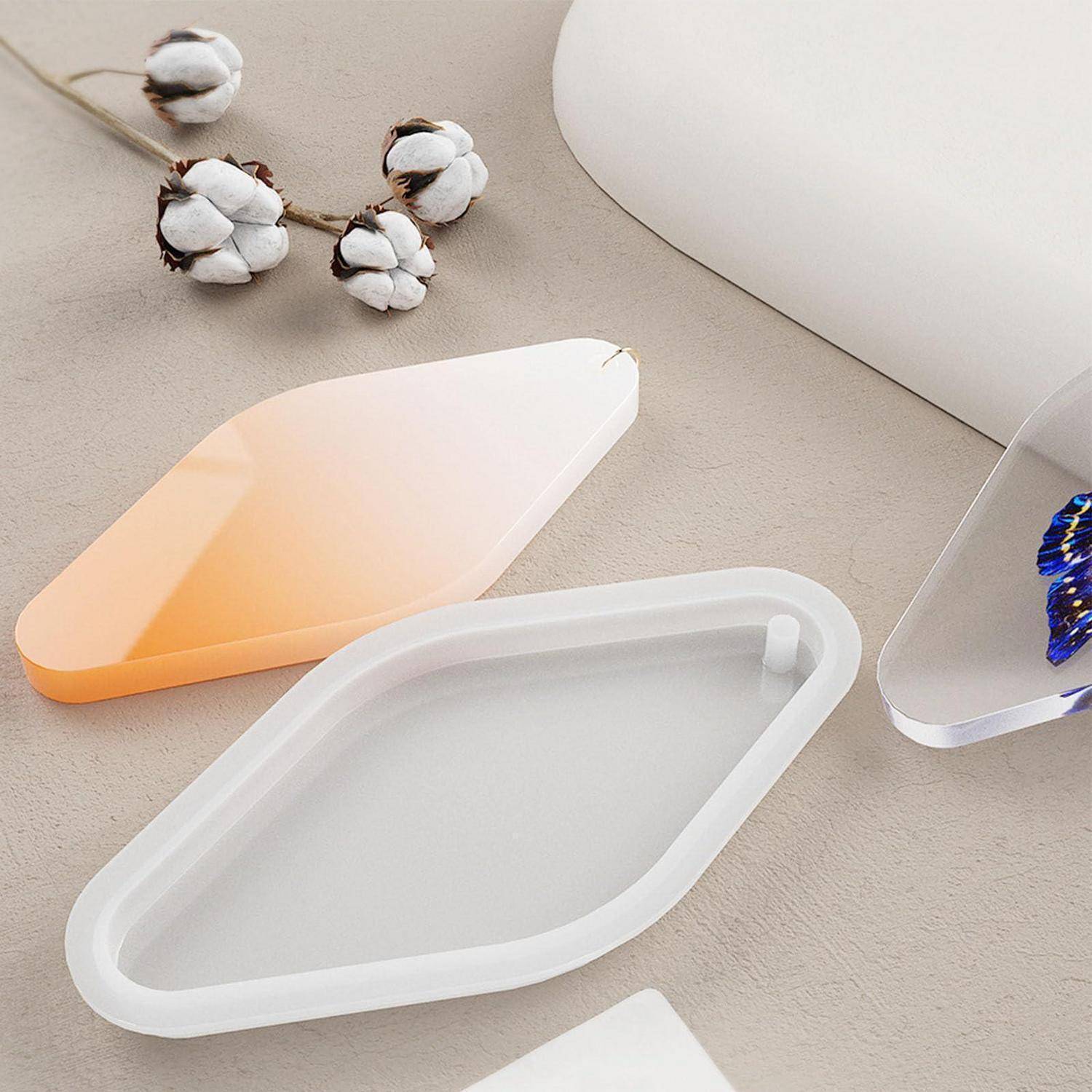 Drei durchscheinende geometrische Silikon-Tabletts auf einer beigen Oberfläche, mit Wattestäbchen in der Nähe, in Pastellorange, Weiß und transparenten Ausführungen.