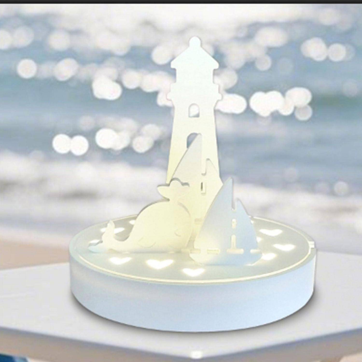 Eine weiße ausgeschnittene Leuchtturm-Pop-up-Lampe sitzt auf einer runden Basis und leuchtet sanft vor einem unscharfen Küstenhintergrund.