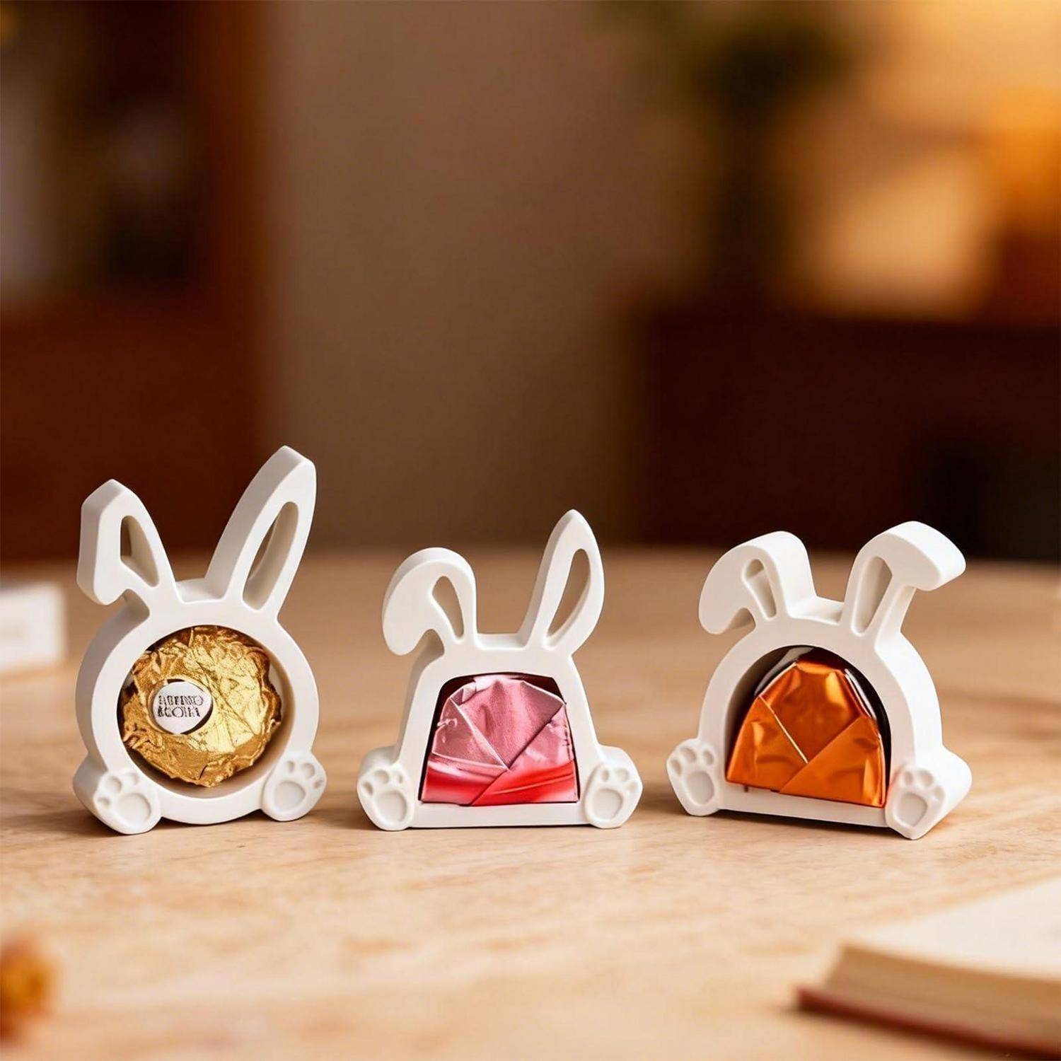Drei hasenförmige Süßigkeitenbehälter auf einem Tisch, von denen jeder eine verpackte Schokolade zeigt: gold, rosa und orange.