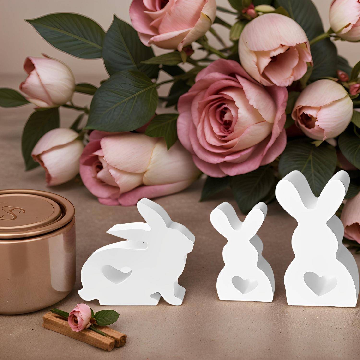Drei weiße kaninchenförmige Dekorationen mit Herzausschnitten neben einem Kupferbehälter und rosa Rosen auf einer Tischplatte.