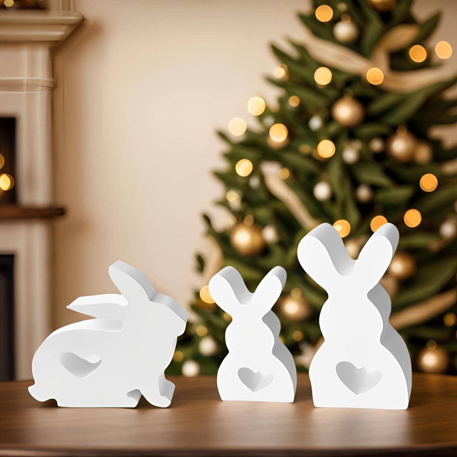 Drei weiße, kaninchenförmige Holzdekorationen auf einem Tisch, mit einem unscharfen beleuchteten Weihnachtsbaum im Hintergrund.