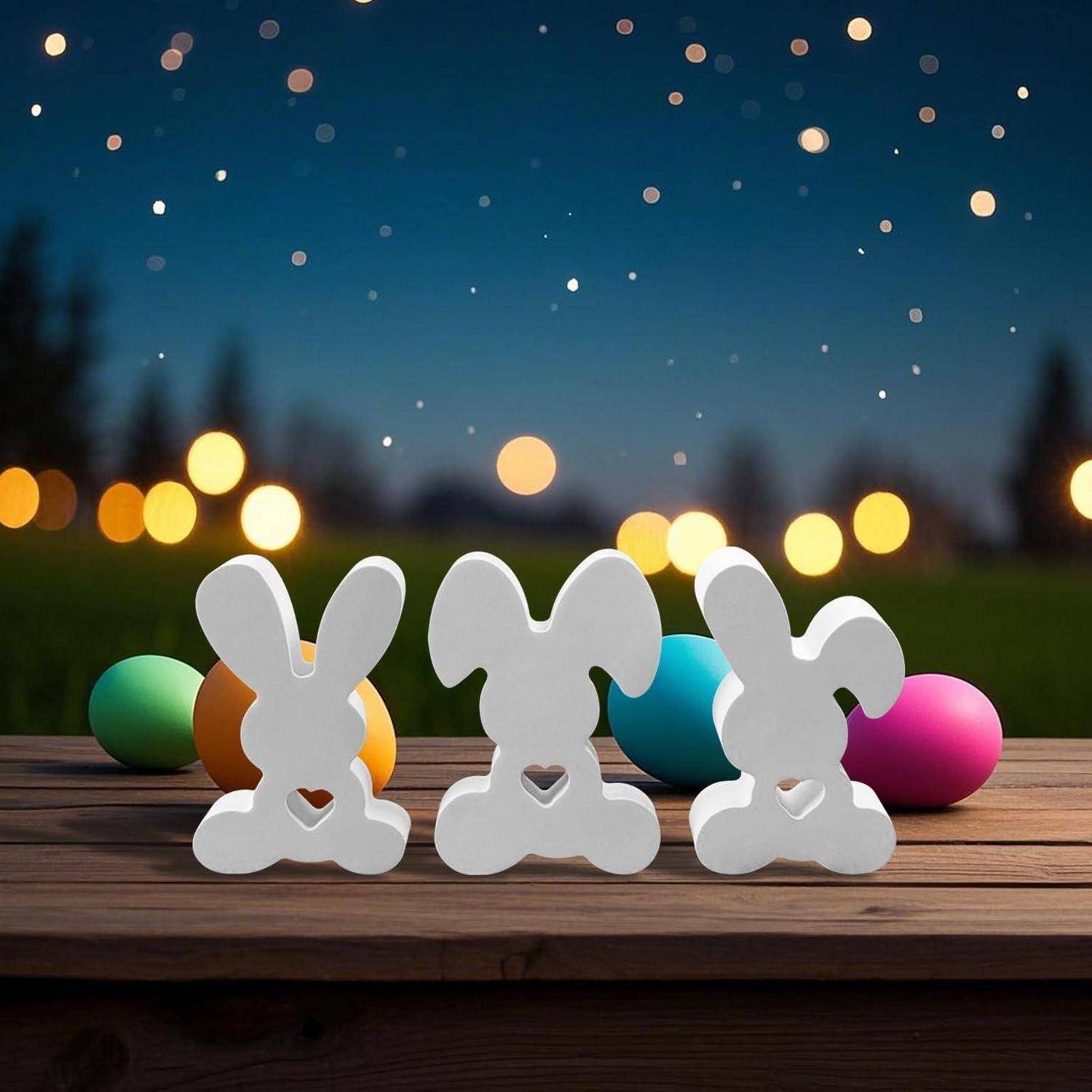 Drei weiße hasenförmige Ausschnitte auf einem Holztisch mit bunten Ostereiern und unscharfem Lichterhintergrund.
