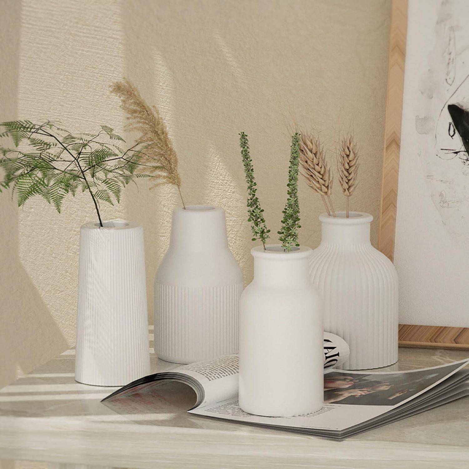 Minimalistische Stillleben mit vier weißen strukturierten Vasen mit Trockenblumen und einem Farn auf einer aufgeschlagenen Zeitschrift vor einer beigen Wand
