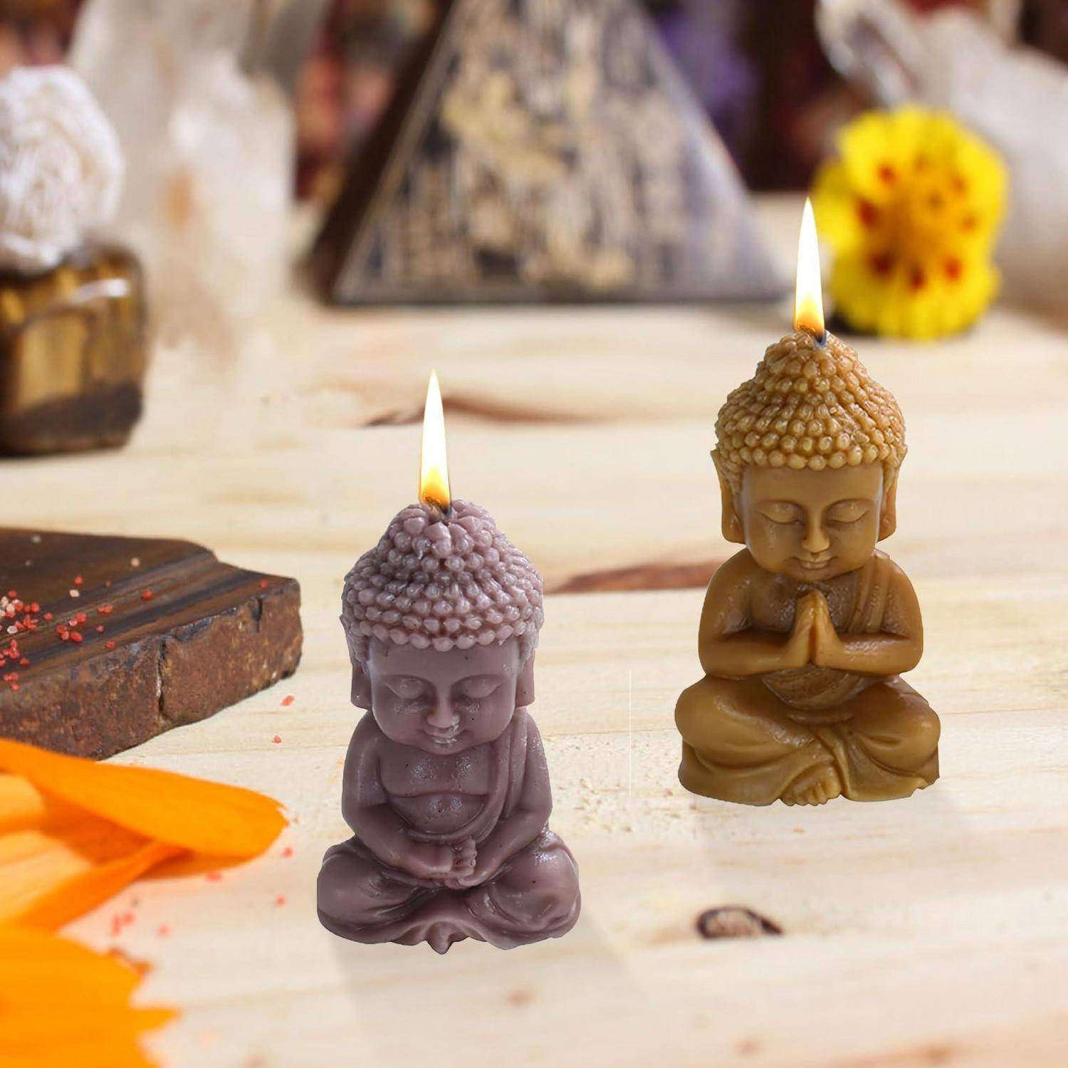 Zwei brennende Buddha-förmige Kerzen stehen auf einer Tischplatte, eine violett und eine golden, mit Räucherstäbchen und Blumen verschwommen im Hintergrund.