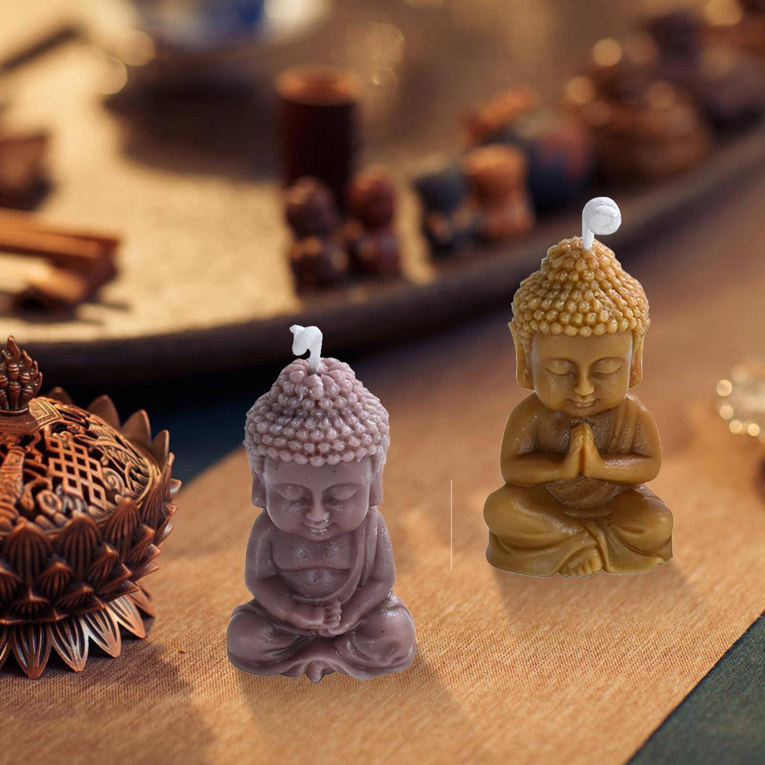 Zwei kleine Buddha-förmige Kerzen, eine Lavendelfarbe und eine golden, stehen auf einem Tisch neben einem dekorativen Kerzenhalter.