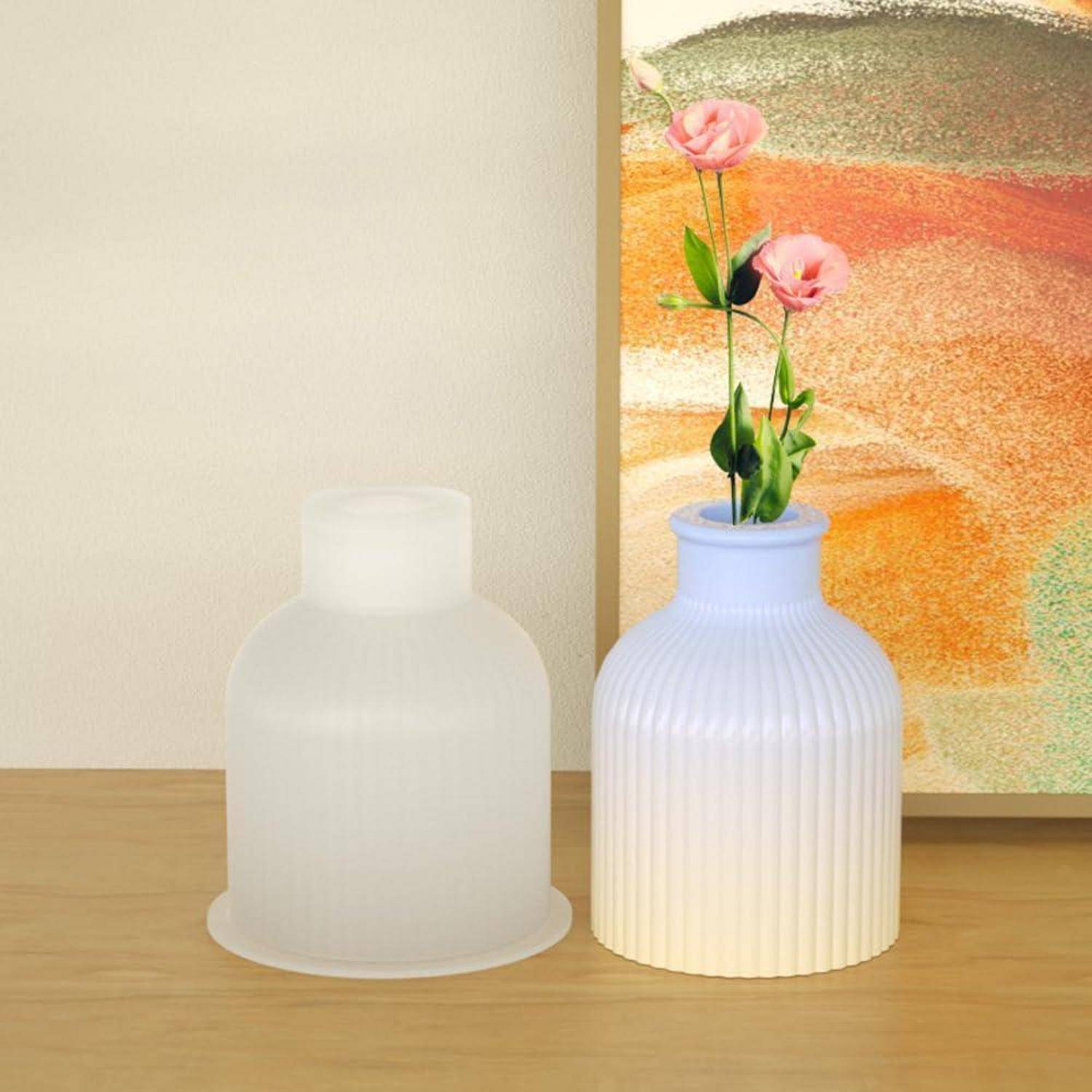 Zwei dekorative Vasen auf einem Holztisch, eine aus mattiertem Glas und leer, die andere weiß geriffelt mit rosa Blumen, vor einem farbenfrohen Gemälde.