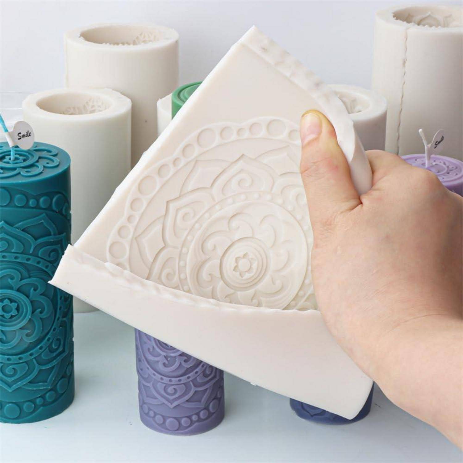 Hand, die eine flexible weiße Silikonform mit einem geprägten Mandala-Design hält, umgeben von mehreren farbigen zylindrischen Formen auf einer weißen Oberfläche.