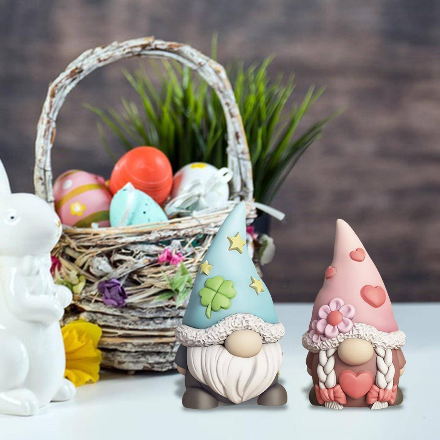 Zwei pastellfarbene Gnoměfigurinen mit Herzen und Blumen sitzen neben einem Korb mit bunten Ostereiern und einer weißen Hasen-Dekoration.
