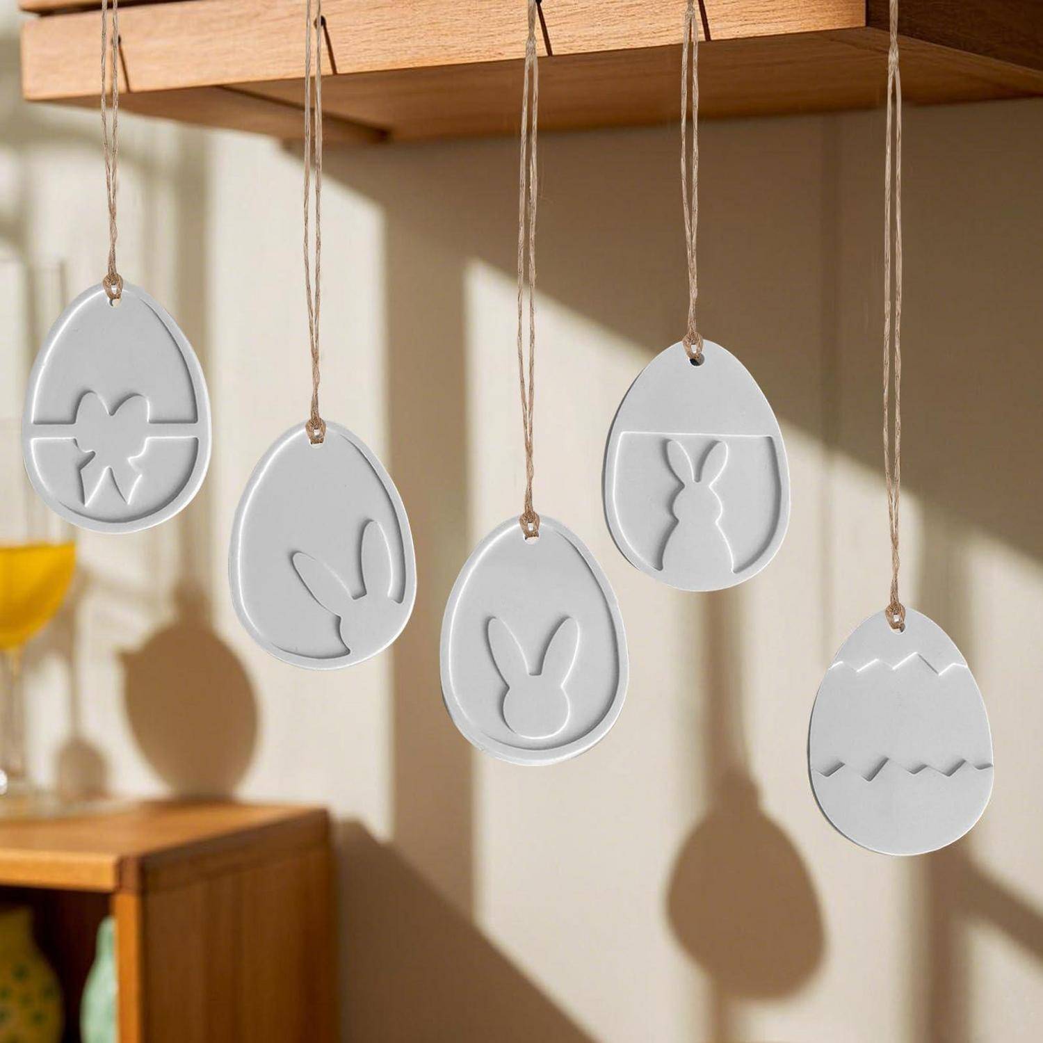 Weiße hängende Oster-Eierverzierungen mit ausgeschnittenen Hasen- und Blumenmustern, aufgehängt an einem Holzregal in einem sonnigen Zimmer.