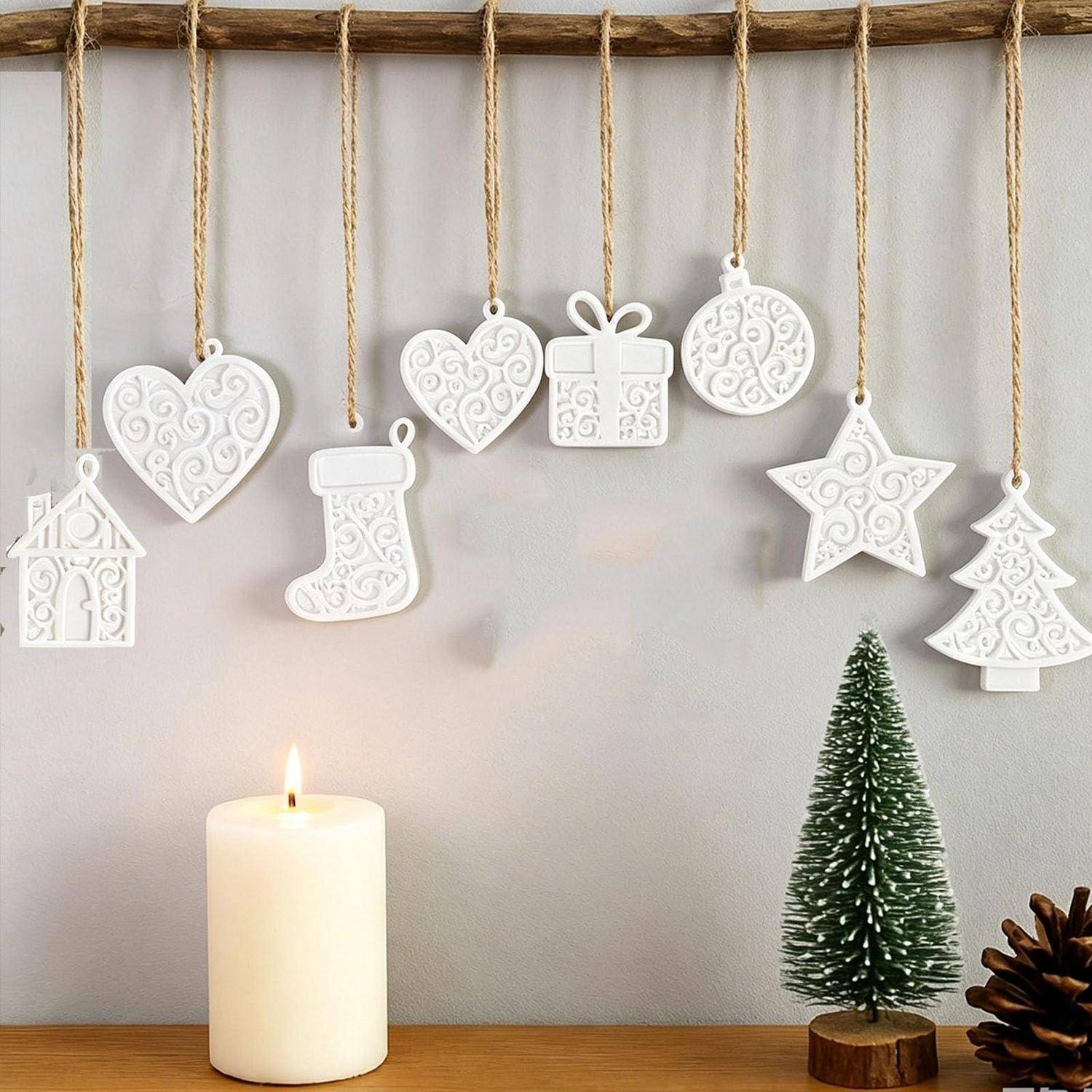 Weiße hängende Weihnachtsverzierungen auf Schnur, darunter Herzen, Geschenk, Strumpf, Stern, Haus und Baum, über einer brennenden Kerze und einem kleinen Tisch-Weihnachtsbaum angeordnet.