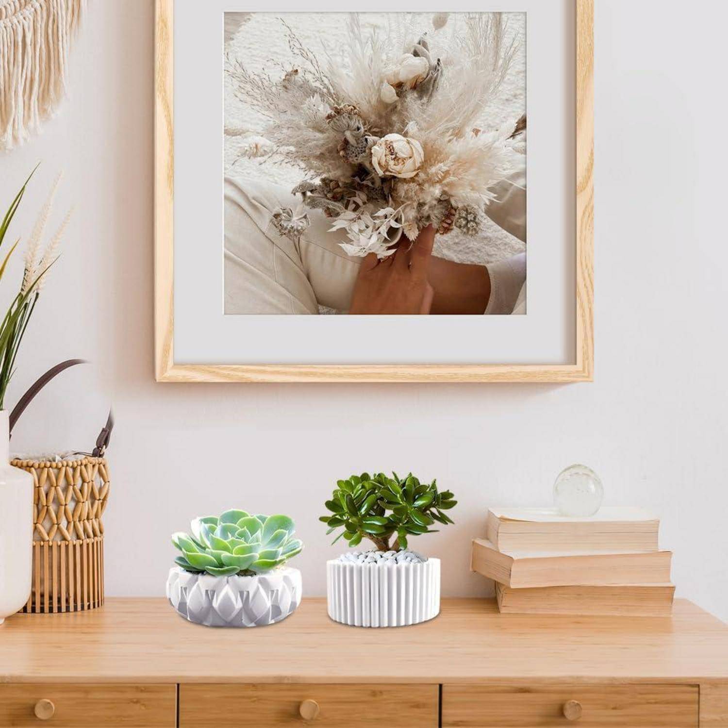 Minimalistisches hölzernes Sideboard mit gerahmter Blumenkunst an der Wand, zwei kleine Töpfe mit Sukkulenten, gestapelte Bücher und Flechtdekoration in einem hellen Zimmer.