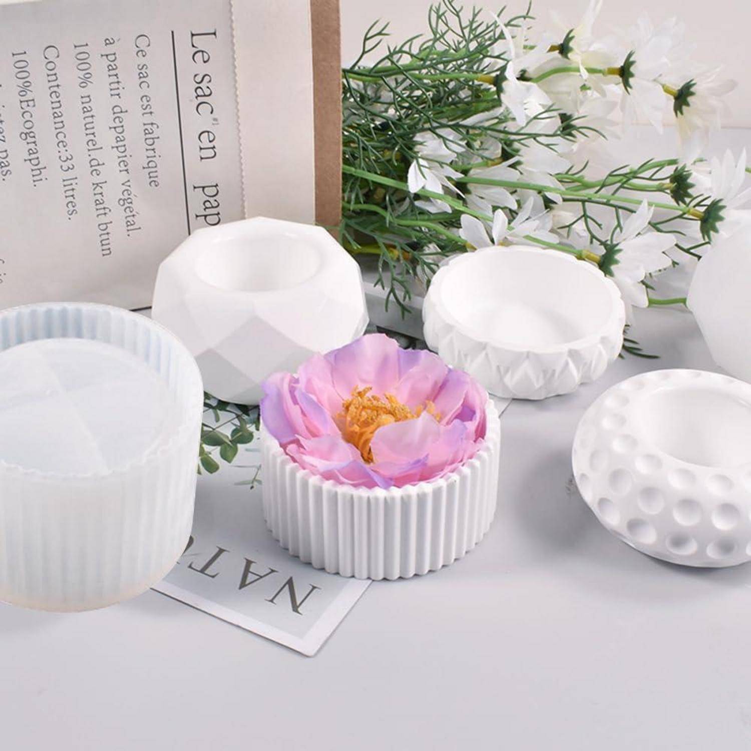 Assortierte weiße dekorative Kerzenhalter und eine rosa Blütenkerze auf einer hellen Oberfläche, mit Grünpflanzen und einer Papiertüte im Hintergrund.