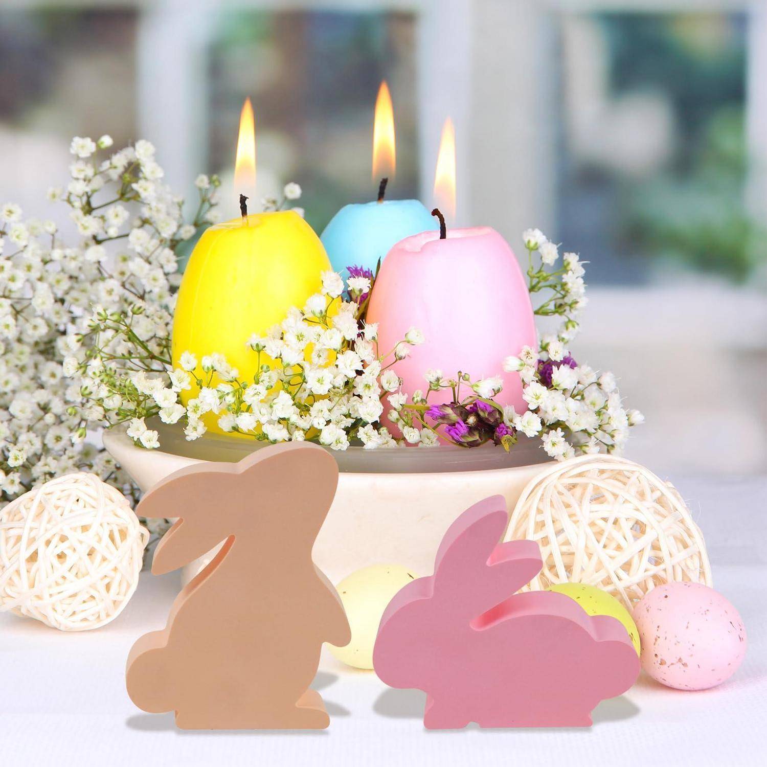 Pastellfarbene Osterdekorationmit drei leuchtenden, eierförmigen Kerzen in Gelb, Blau und Rosa, umgeben von Blumen und dekorativen Hasenschnitt-Ornamenten sowie geflochtenen Kugeln.