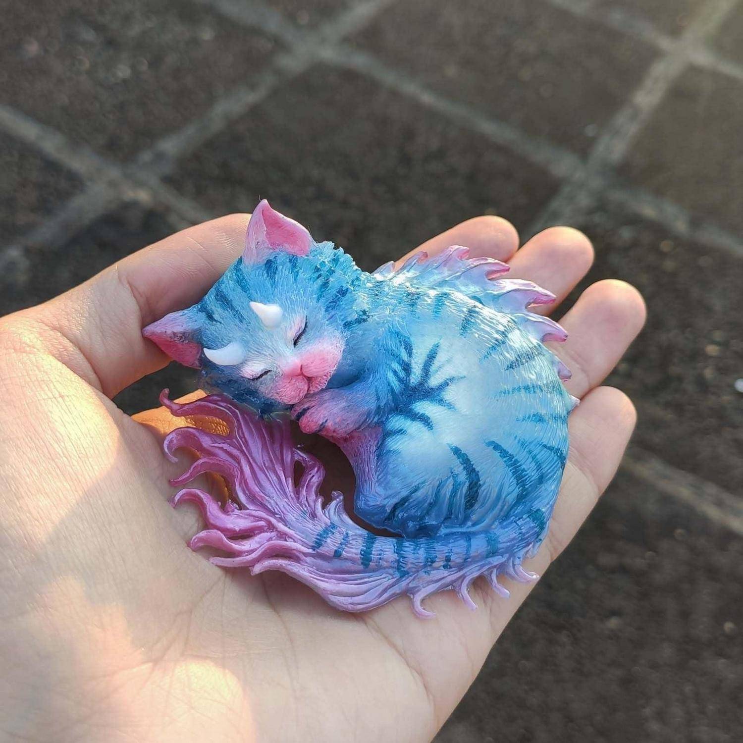 Eine kleine bunte Katzenfigur, die schläft, in Blau und Rosa, zusammengerollt in der offenen Handfläche einer Person.