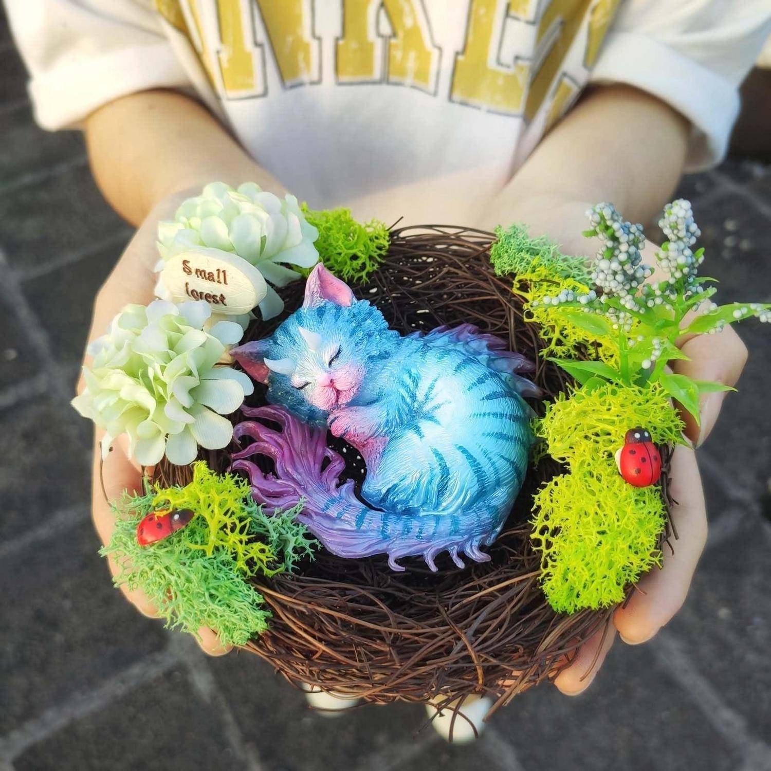 Hände halten ein dekoratives Vogelnest mit einer blauvioletten schlafenden Katzenfigur, Blumen, Moos und kleinen roten Marienkäfern.