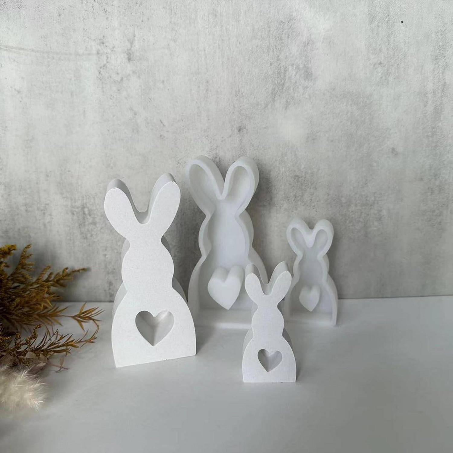 Weiße dekorative Hasenfiguren in verschiedenen Größen, jeweils mit herzförmigen Ausschnitten, auf einer Tischplatte vor einer grau strukturierten Wand angeordnet.