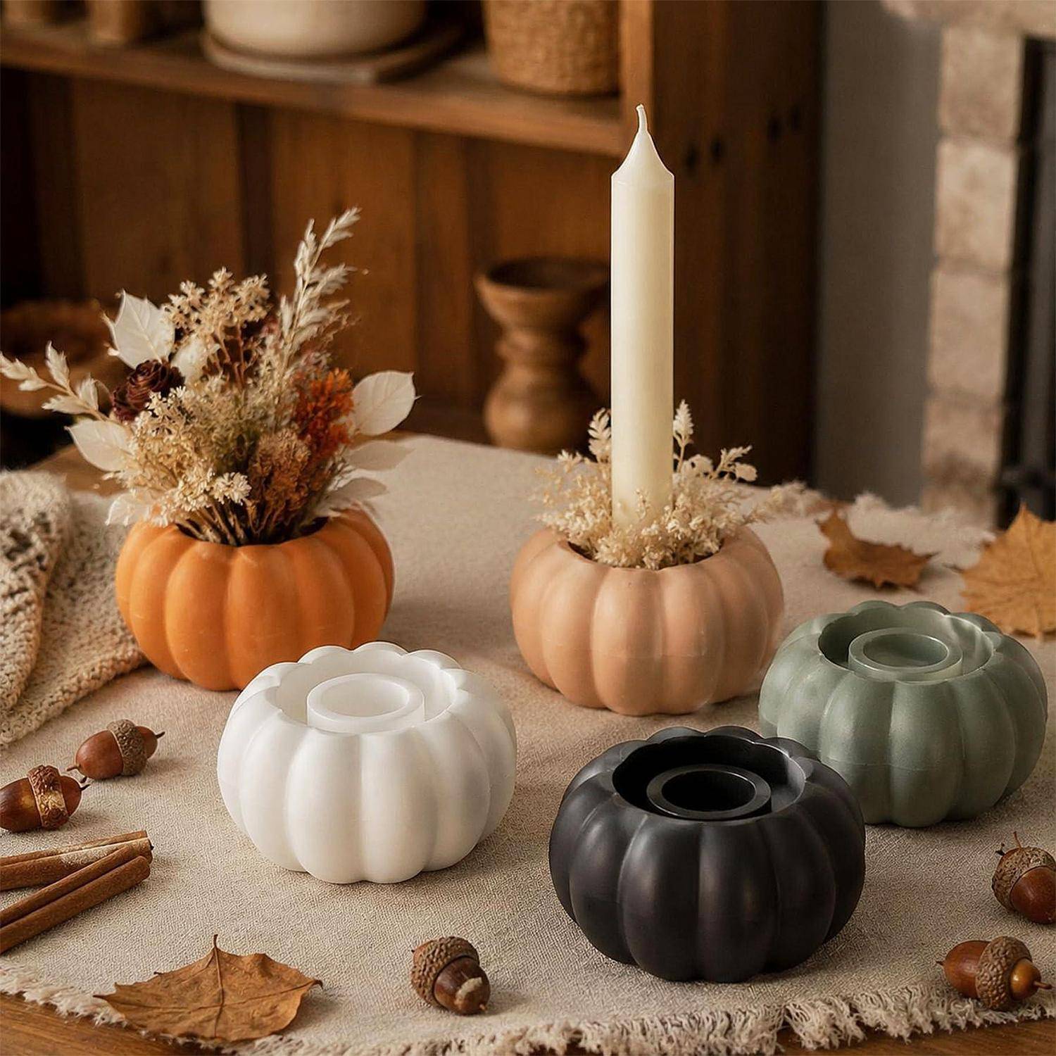 Herbstliche Tischdekoration mit kürbisförmigen Kerzenhaltern in Orange, Weiß, Beige, Grün und Schwarz auf einem Leinentuch, umgeben von getrockneten Blättern und Eicheln.