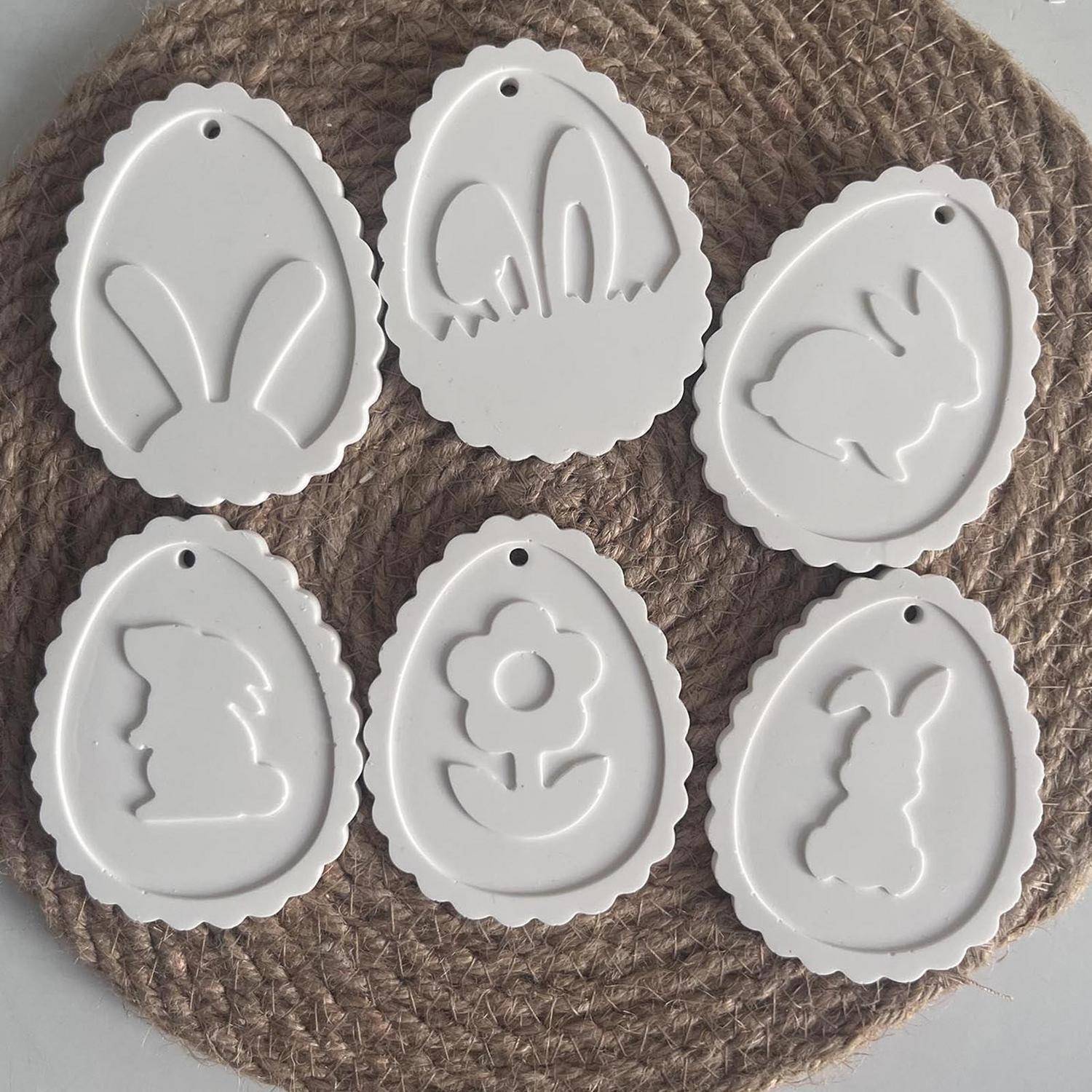 Sechs weiße Oster-Hängornamente mit gewellten Kanten und ausgeschnittenen Designs von Hasenohren, Hase, Blume und Kaninchen, angeordnet auf einer gewebten Matte.