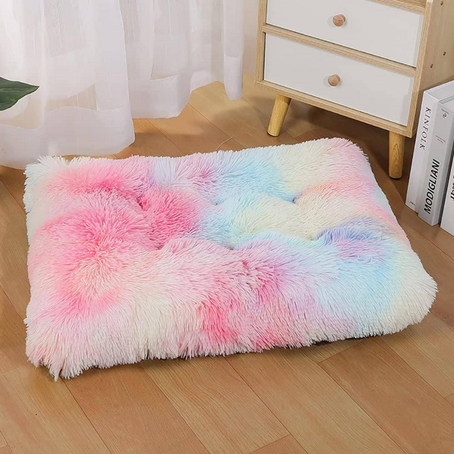 Flauschiges Bodenkissen oder Haustieratte in Pastellrosa, Blau und Weiß auf einem Holzboden neben einer kleinen Kommode und Büchern.