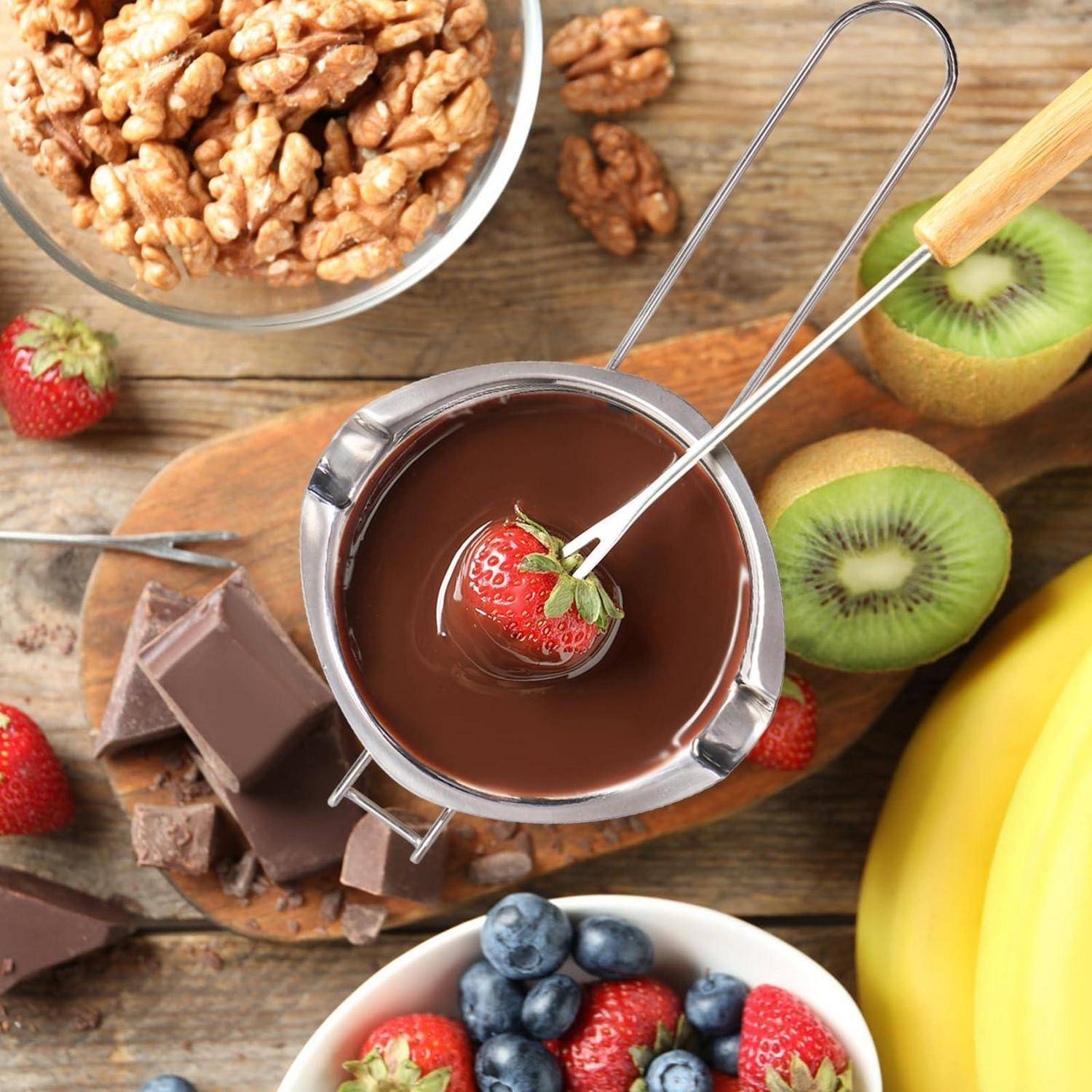 Schokoladenfondue in einem Metalltopf mit einer Erdbeere auf einer Gabel, umgeben von Obst, Banane, Kiwi, Walnüssen und Schokoladenstücken auf einem Holztisch.