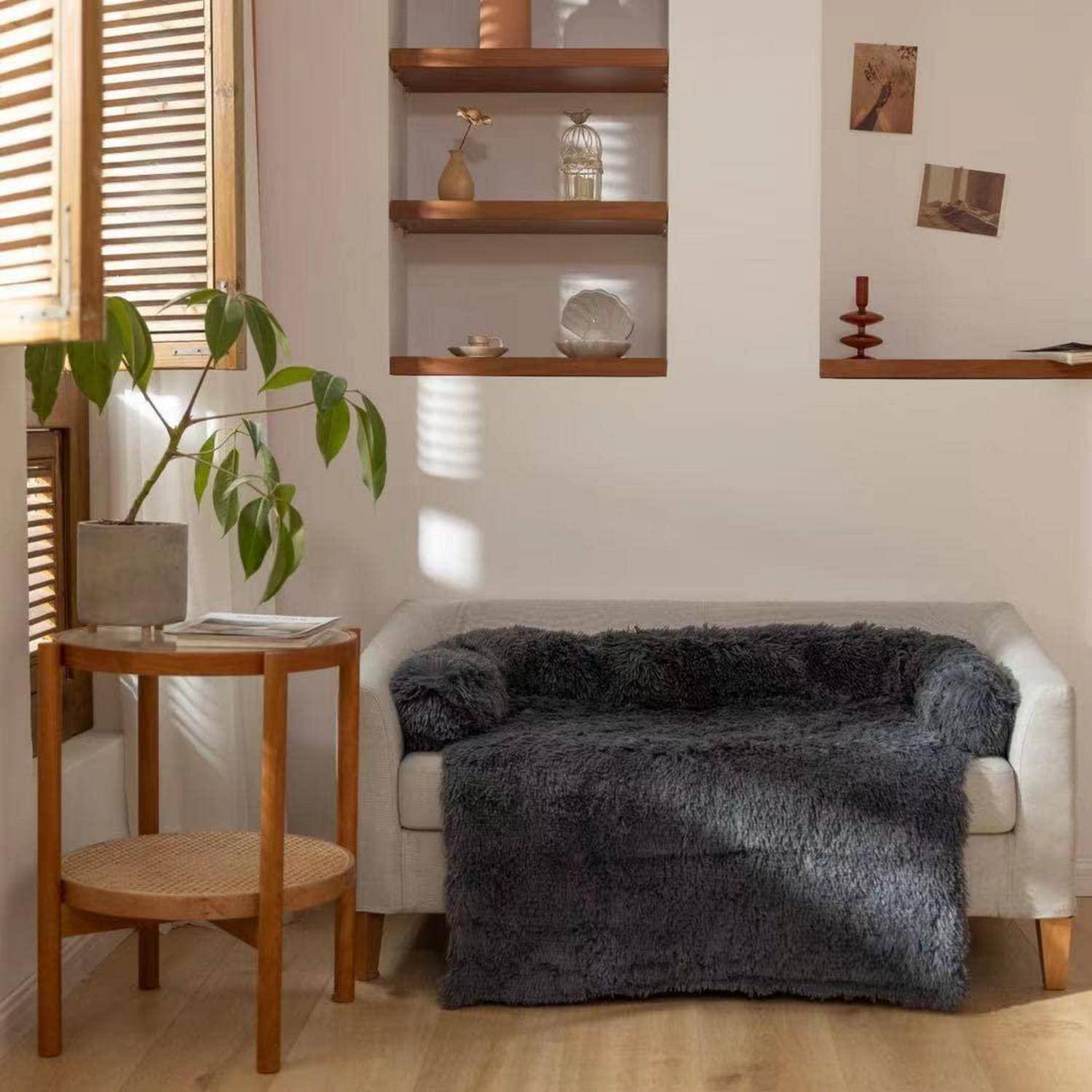 Gemütliches Wohnzimmer mit einem weißen Sofa, das mit einer dunkelgrauen Wurfdecke drapiert ist, einem kleinen hölzernen Beistelltisch mit einer Pflanze und Wandregalen mit Dekoration im warmen Sonnenlicht.