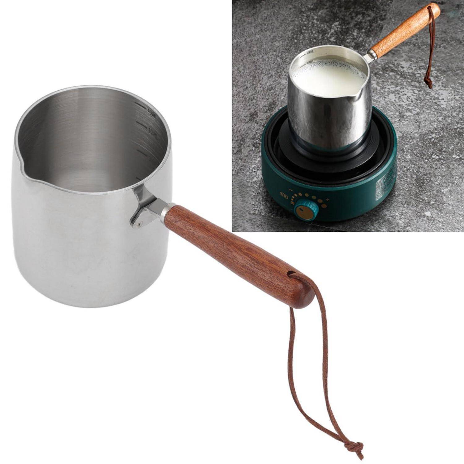 Edelstahlkännchen zum Aufschäumen von Milch mit Holzgriff, allein gezeigt und auf einer kleinen elektrischen Kochplatte mit Milch, die darin erhitzt wird.