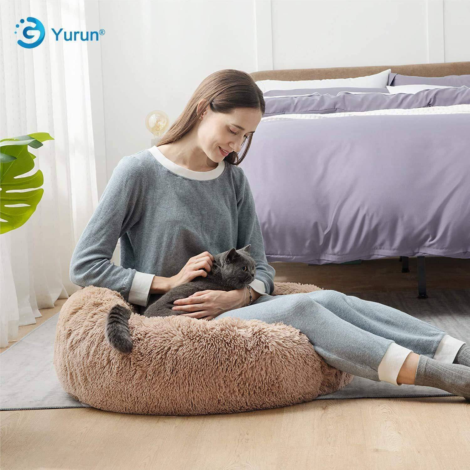 Frau, die auf einem flauschigen Haustierbett sitzt, hält sanft eine Katze in einem Schlafzimmer; das Yurun-Logo erscheint oben links.