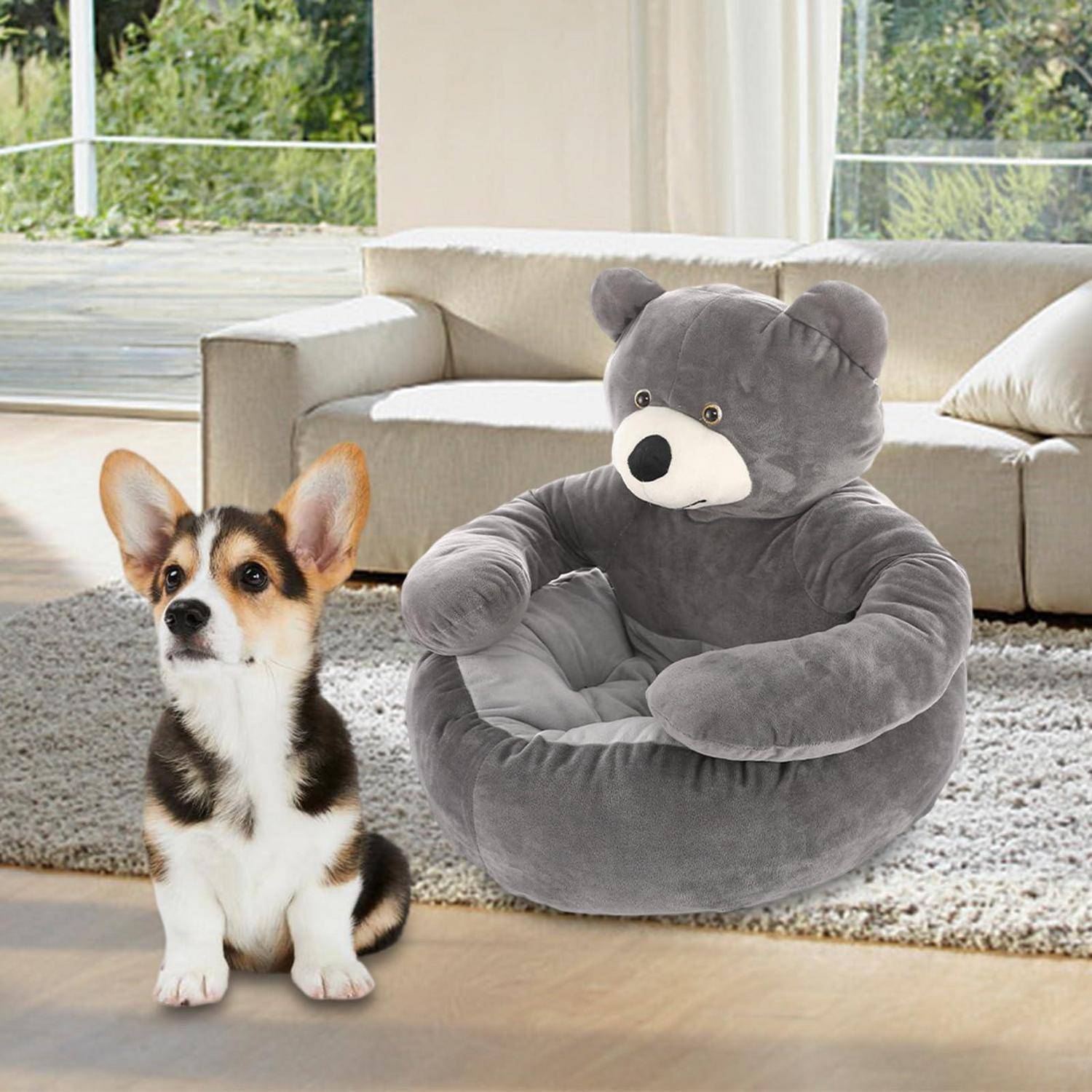 Ein Corgi-Welpe sitzt neben einem grauen Teddybär-Haustierbett in einem hellen Wohnzimmer mit einem Sofa und einem Teppich.