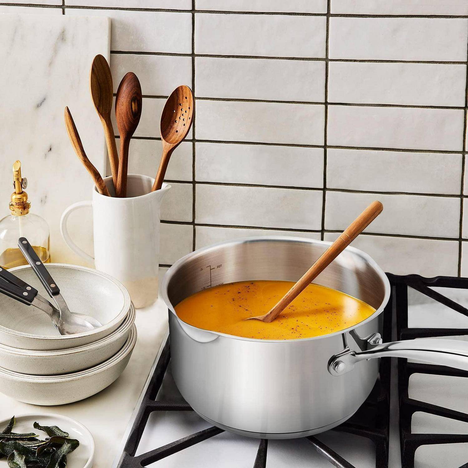 Edelstahlpfanne mit orangefarbener Suppe, die auf einem Kochfeld köchelt, mit Holzlöffel und Küchenutensilien in der Nähe auf einer gefliesten Arbeitsplatte mit Fliesenspiegel.