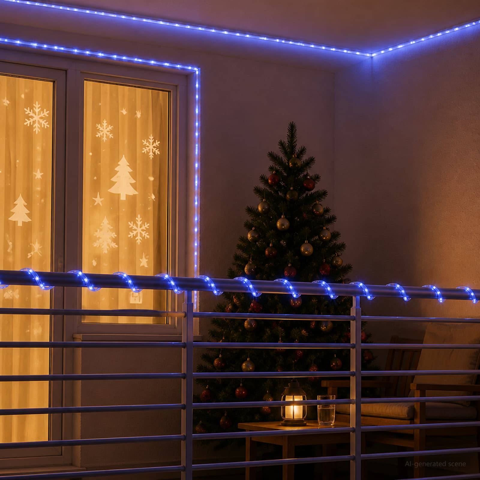 Kleiner Balkon weihnachtlich dekoriert, mit blauen Lichterketten, einem beleuchteten Baum und einer Laterne neben einem Fenster mit Schneeflocken-Vorhängen.