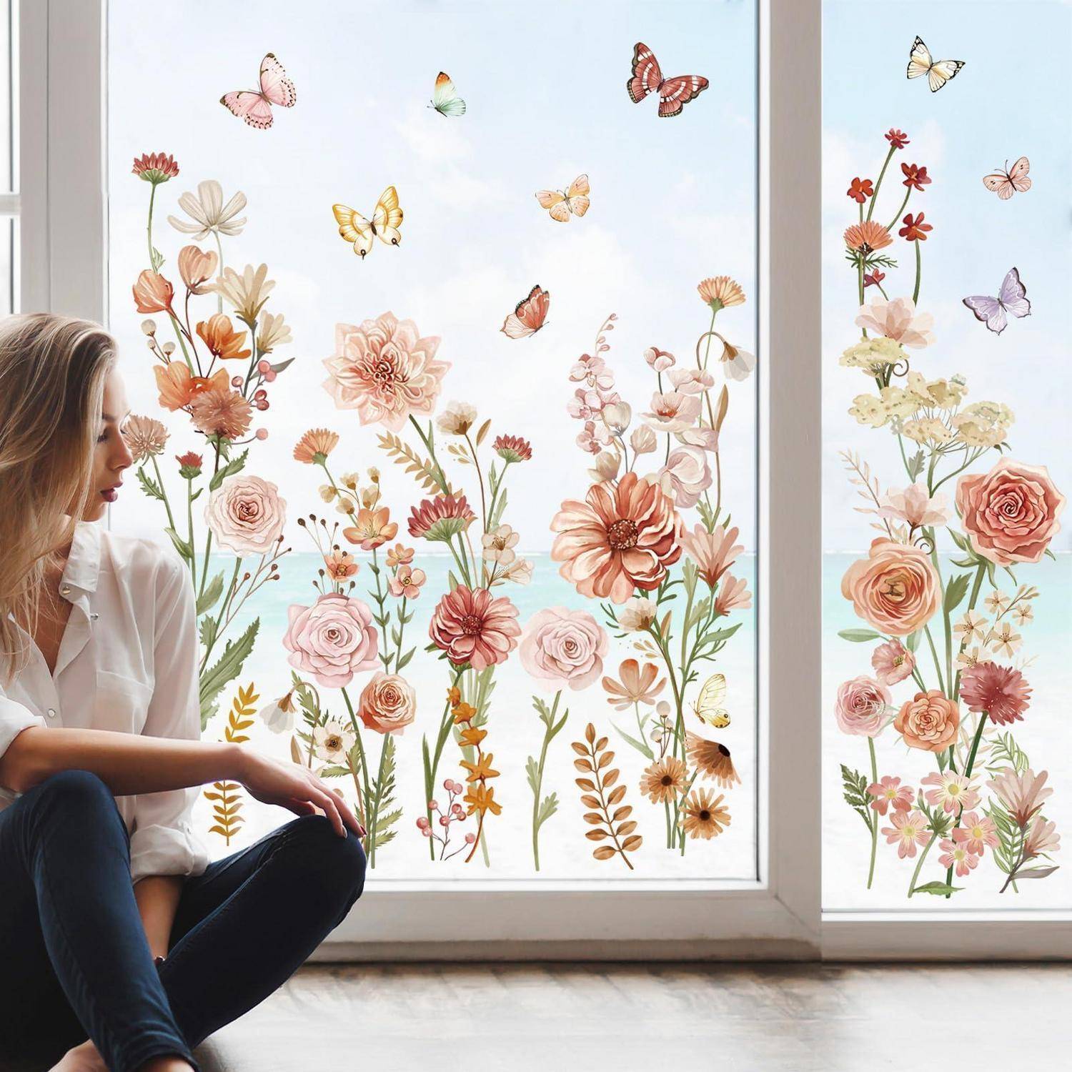 Eine Frau sitzt neben einem Fenster, umgeben von floralem und Schmetterlingskunstwerk an der Wand, wodurch eine helle, frühlingshaft gestaltete Inneneinrichtungsszene entsteht.