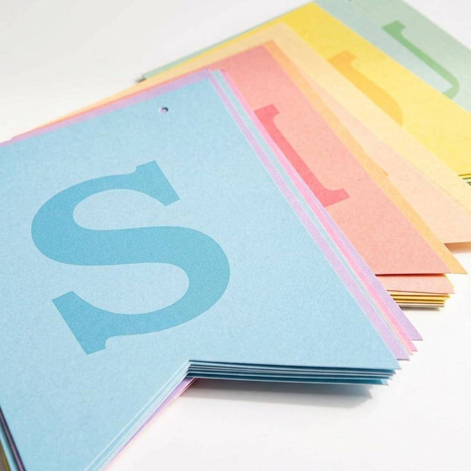 Stapel von pastellfarbenen Alphabet-Lernkarten mit einer großen blauen 'S'-Karte vorne und anderen Buchstaben dahinter auf weißem Hintergrund.