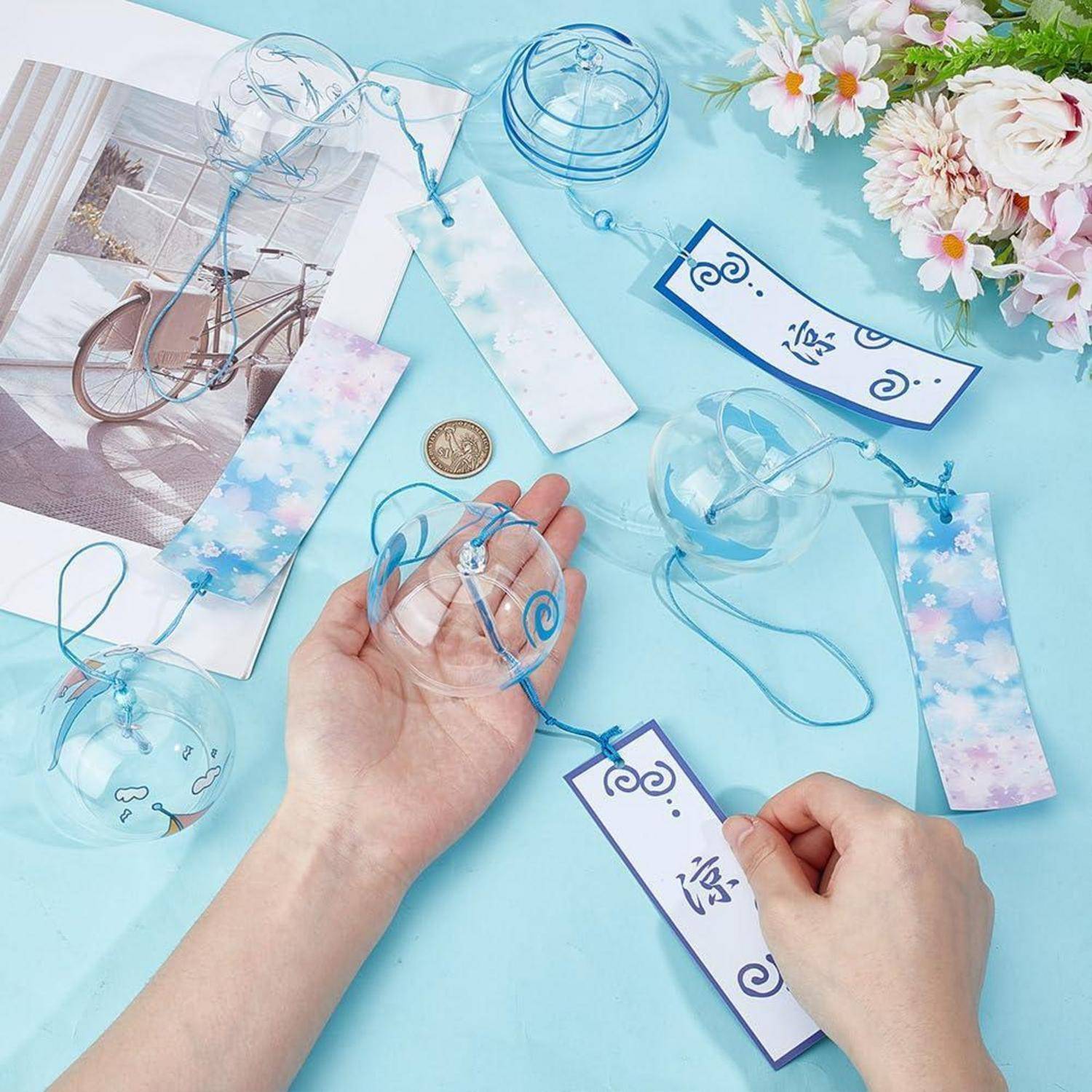 Hände, die ein blaues Band an einem klaren Ornament binden, mit Papierkettchen, Blumen und Fotografie-ähnlichen Bastelmaterialien auf hellblauem Hintergrund.