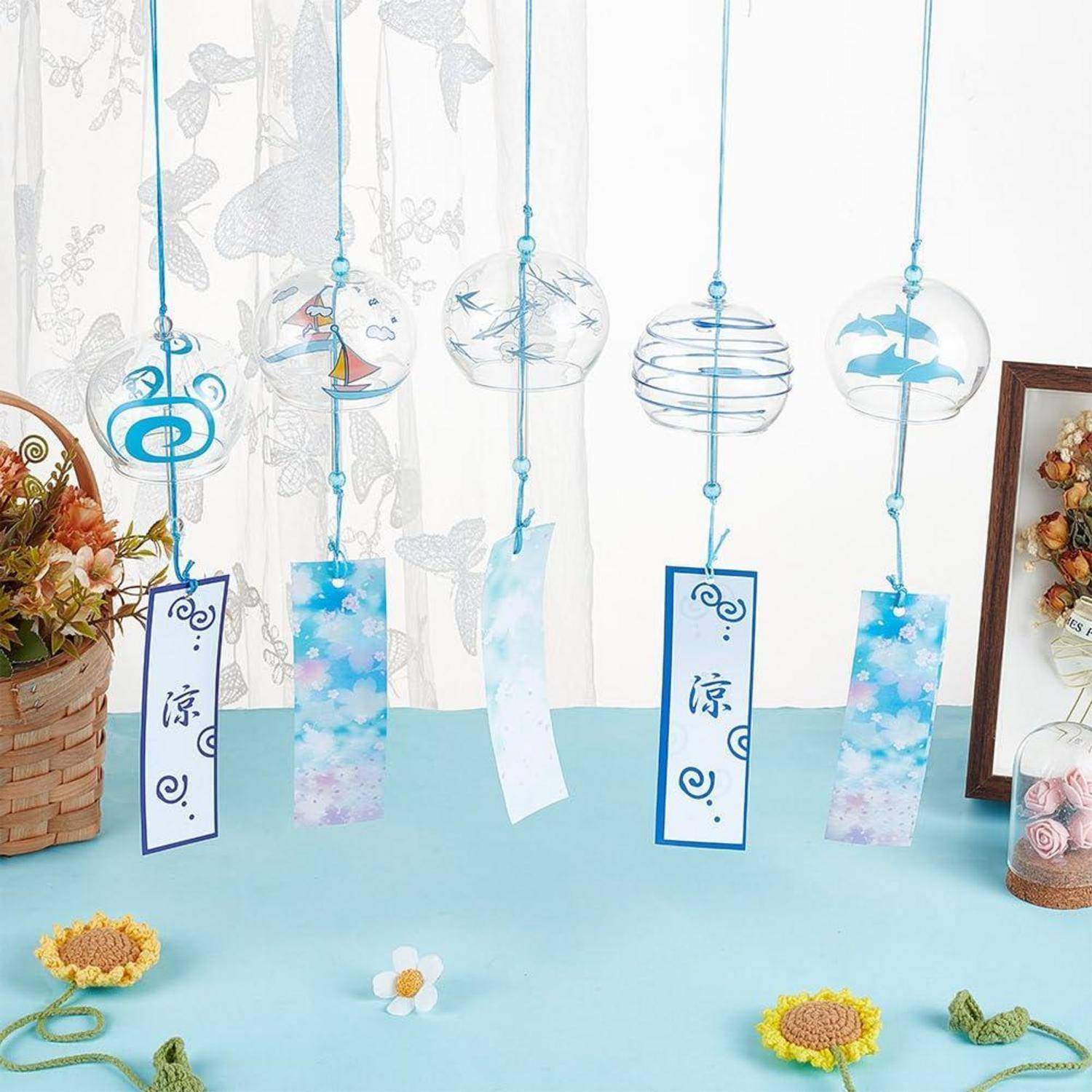 Fünf hängende sommerliche Windspiele und Papieretikett schmücken einen blauen Tisch, mit Blumenakzenten und einem Blumenkorb daneben.