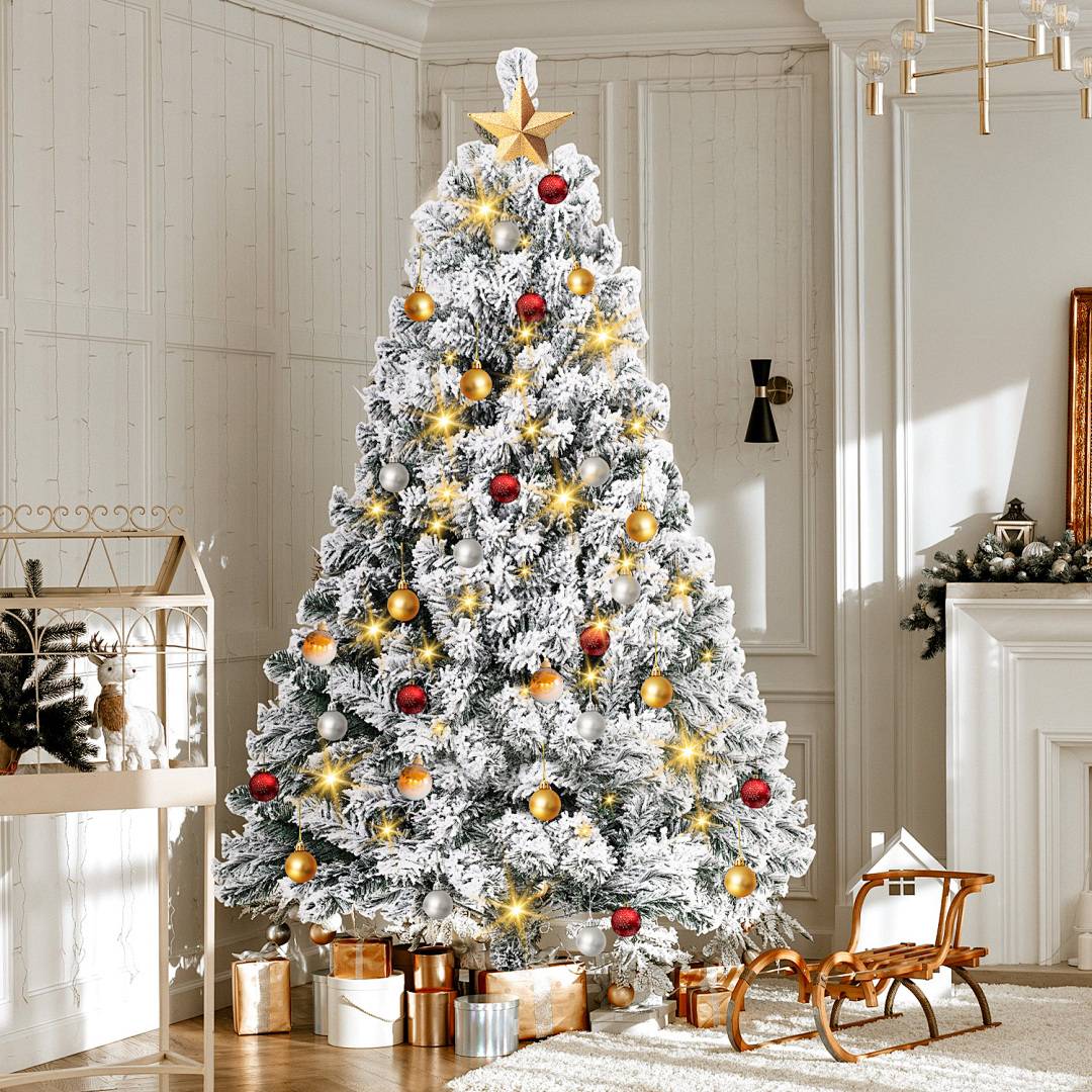 Ein schneebedeckter Weihnachtsbaum mit goldenem Baumspitze, Lichtern und roten und goldenen Kugeln steht in einem hell dekorierten Wohnzimmer.