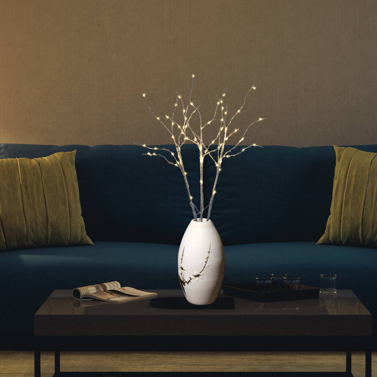 Modernes Wohnzimmer mit dunkelem Sofa, grünen Kissen und einem Couchtisch, auf dem ein aufgeschlagenes Buch, ein Glas, ein Tablett und eine leuchtende weiße Vase mit Astlichtern stehen.