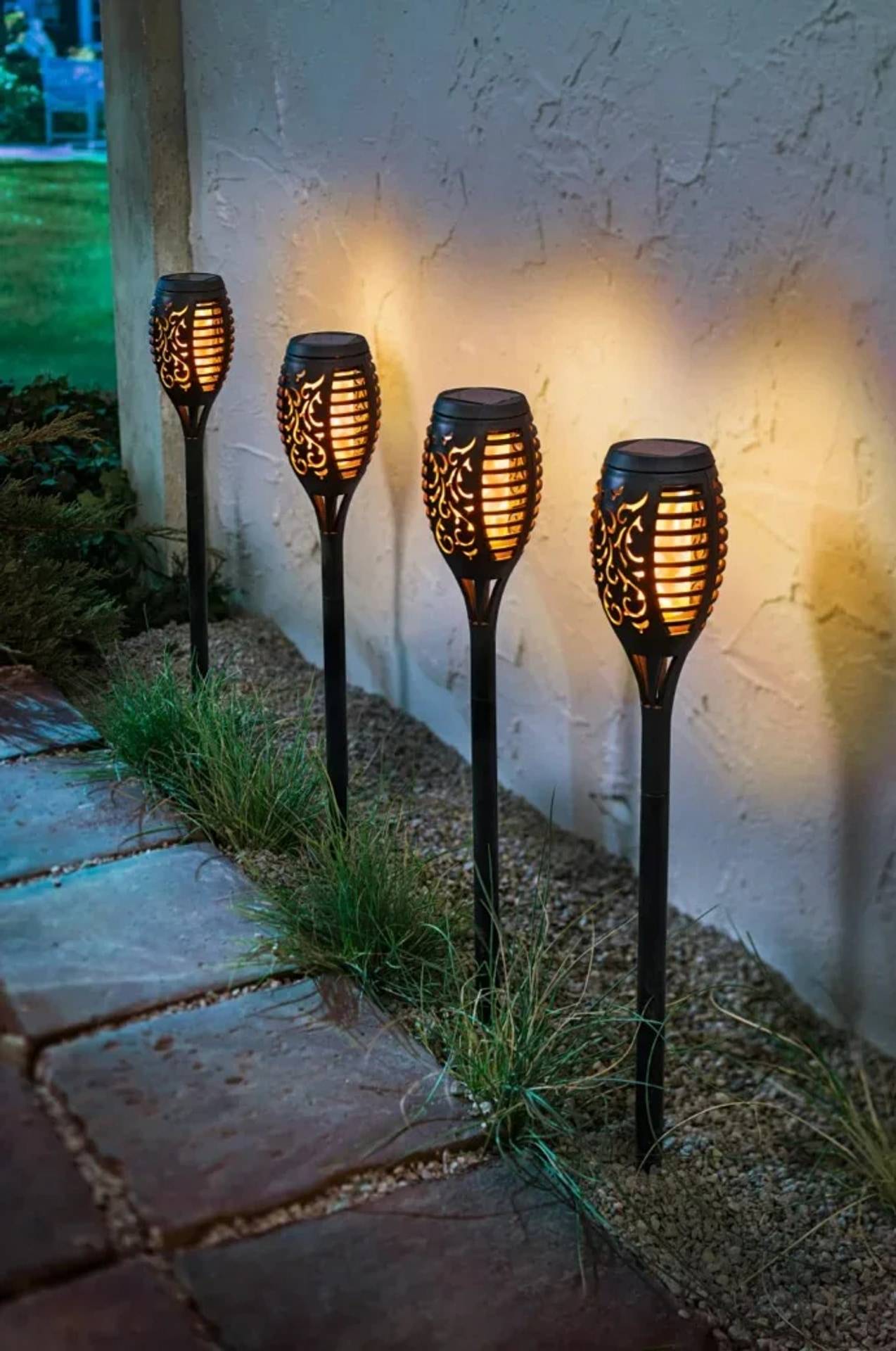 Vier Gartenlichtleuchten im Tiki-Stil leuchten neben einer Stuckwand entlang eines Steinweges in der Dämmerung.