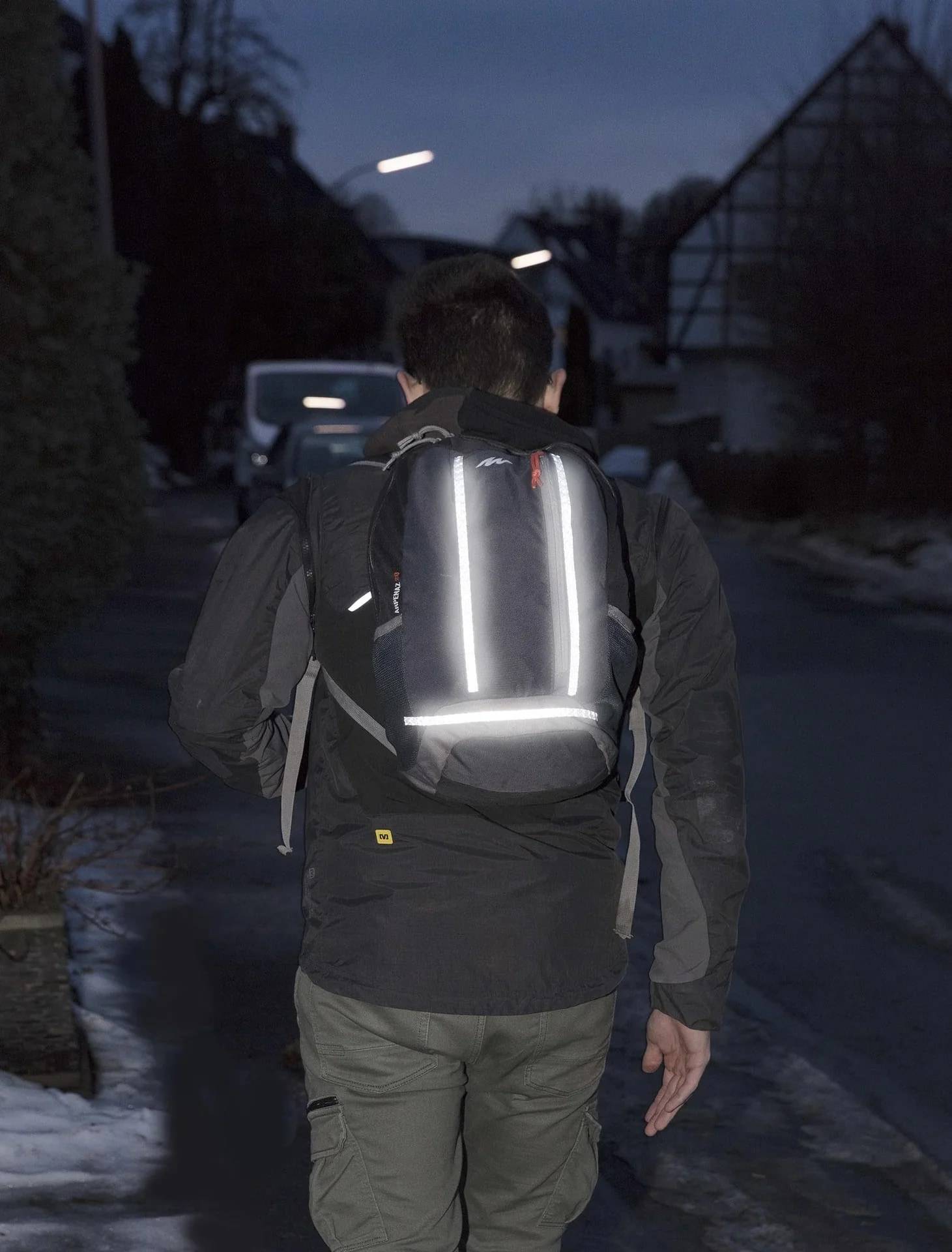 Person, die nachts auf einer dunklen Straße weggeht und einen Rucksack mit reflektierenden Streifen für bessere Sichtbarkeit trägt.