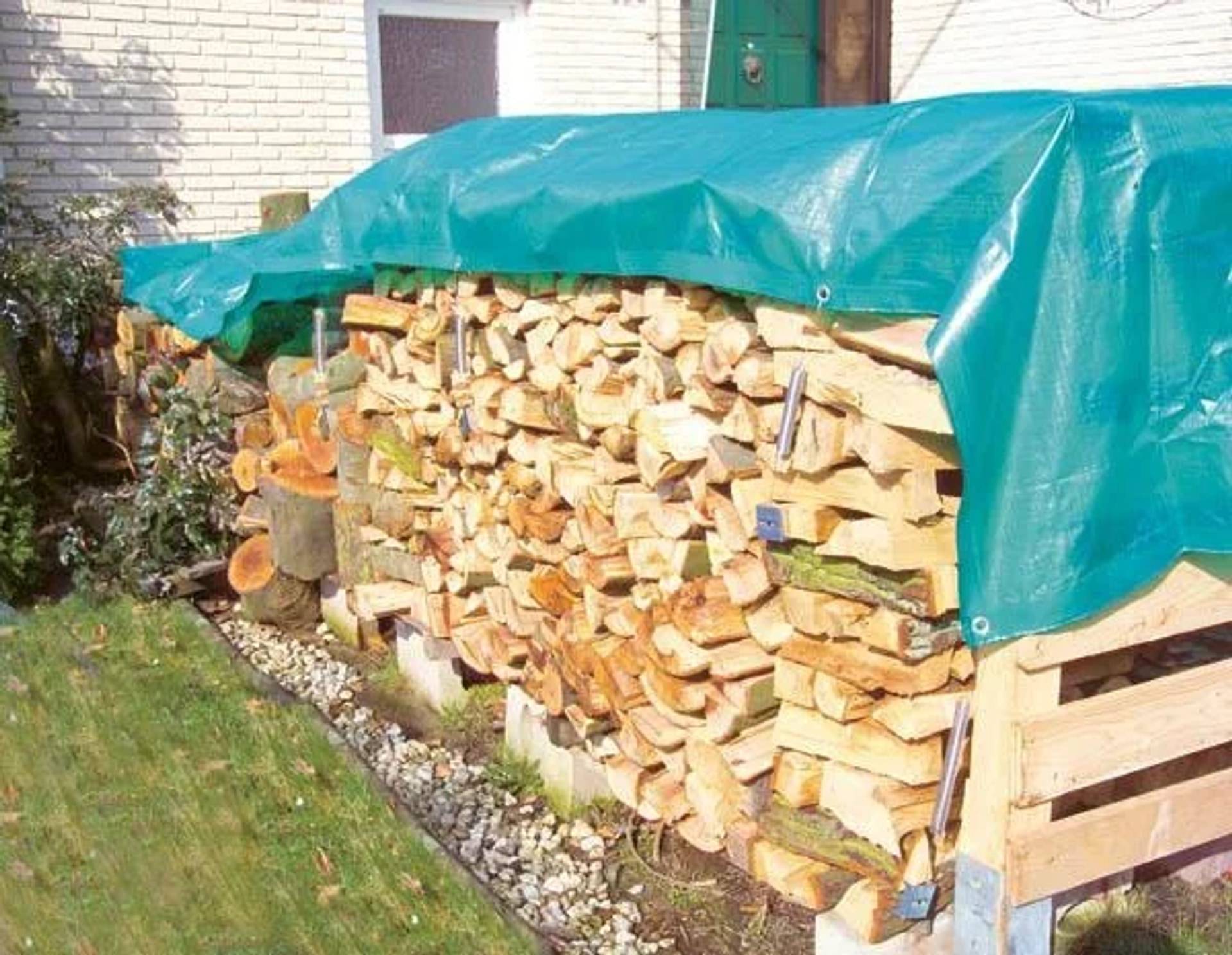 Stapel von gespaltenem Brennholz, das mit einer grünen Plane neben einem Haus auf einem Hof abgedeckt ist.