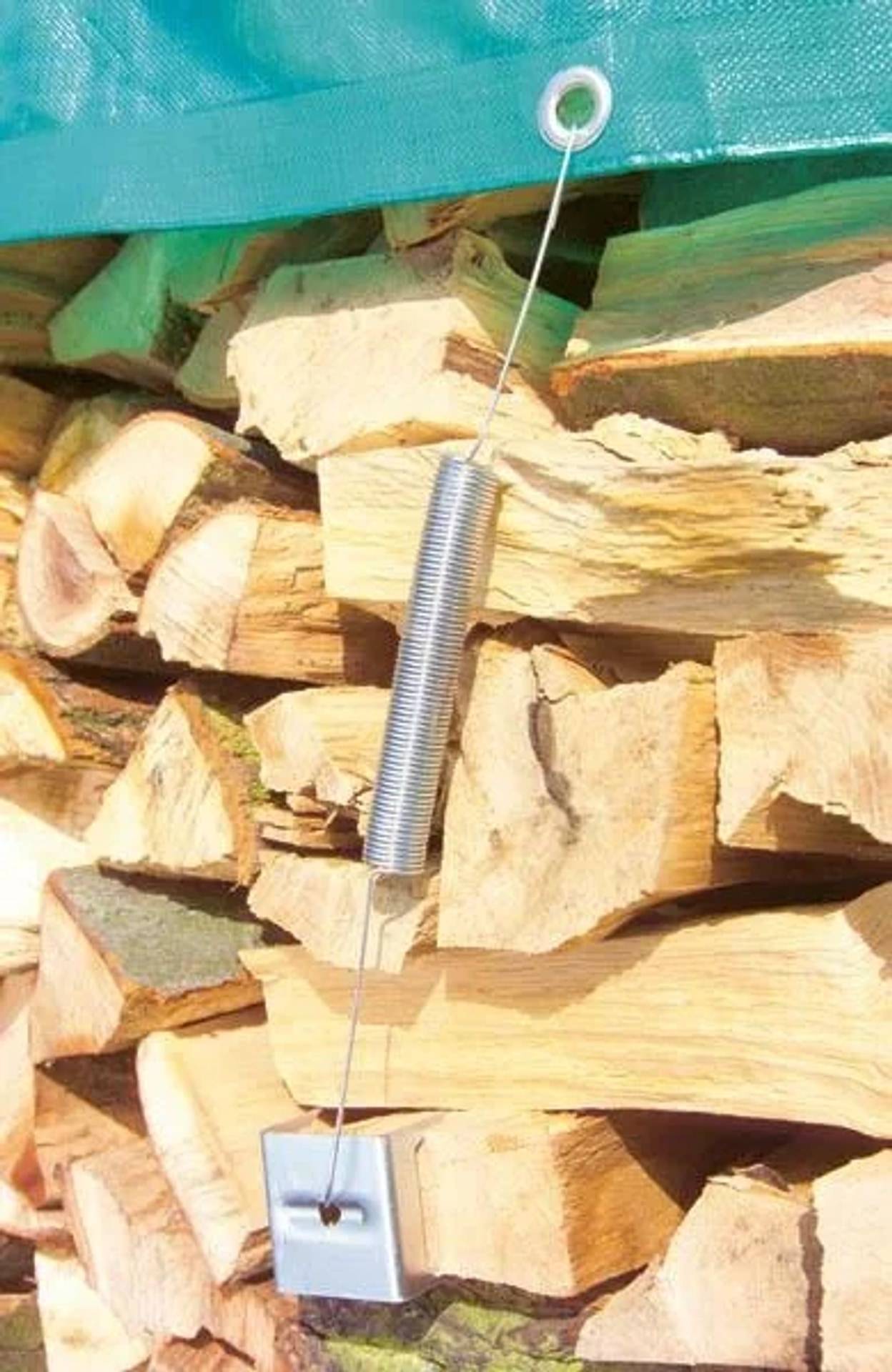Federbelasteter Draht und Anker, die einen gestapelten Holzstapel unter einer grünen Plane im Freien sichern.