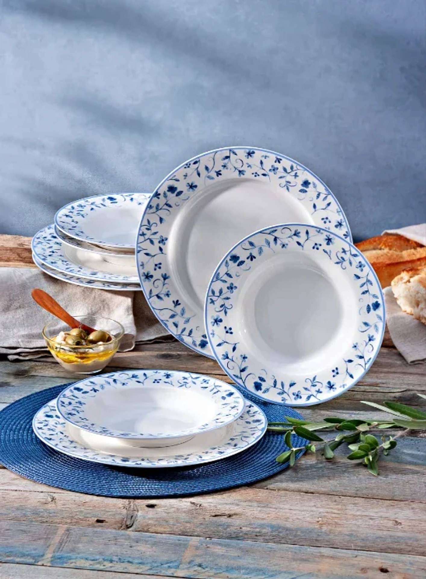 Stapel von weißen Keramiktellern und Schüsseln mit blauen Blumenrändern auf einem rustikalen Holztisch, stylisiert mit Oliven, Brot und einer blauen Platzdecke