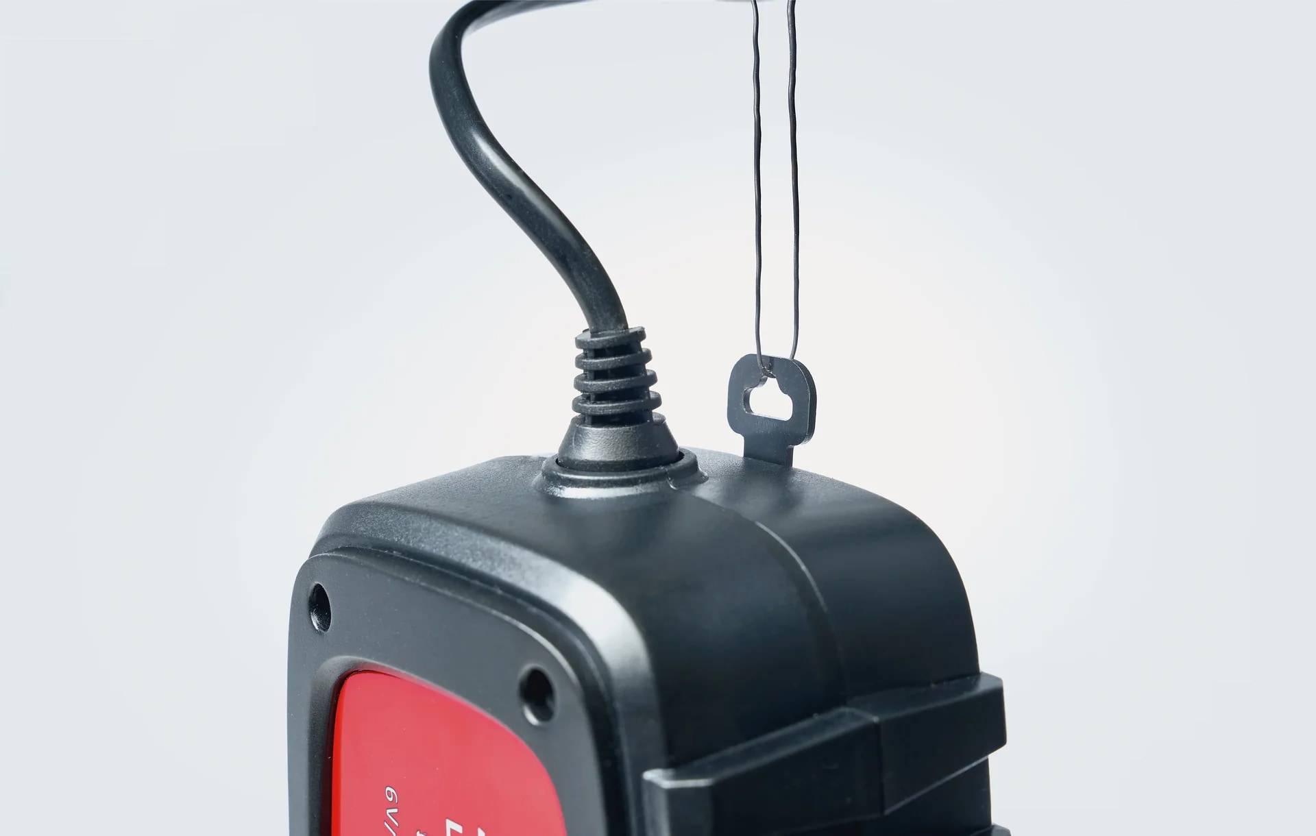 Nahaufnahme eines schwarz-roten hängenden Elektrogeräts mit Kabel und Aufhängungsschlaufe vor einem einfachen, hellen Hintergrund.