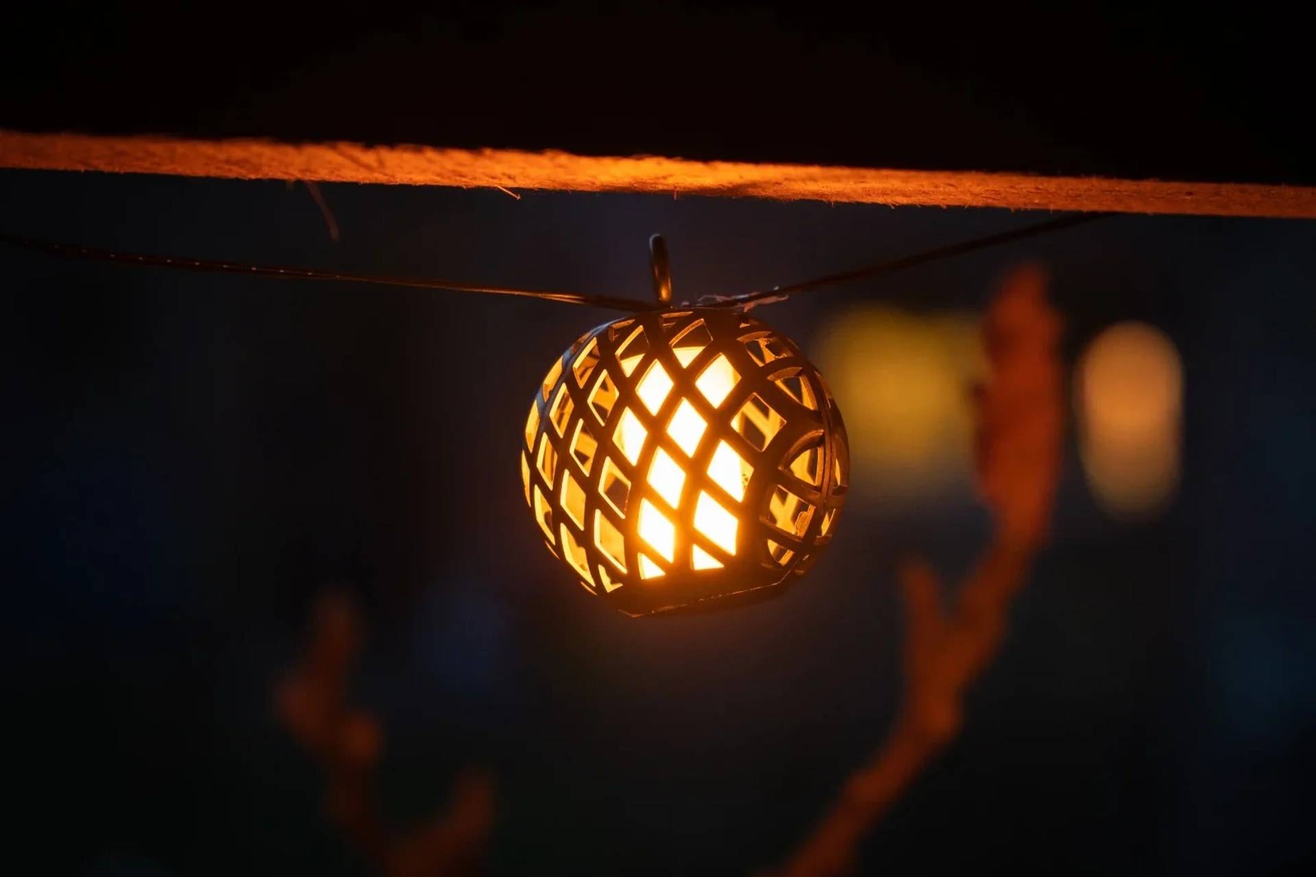 Eine leuchtende geflochtene Laterne hängt an einem Seil in einer dunklen Umgebung und wirft warmes orangefarbenes Licht.