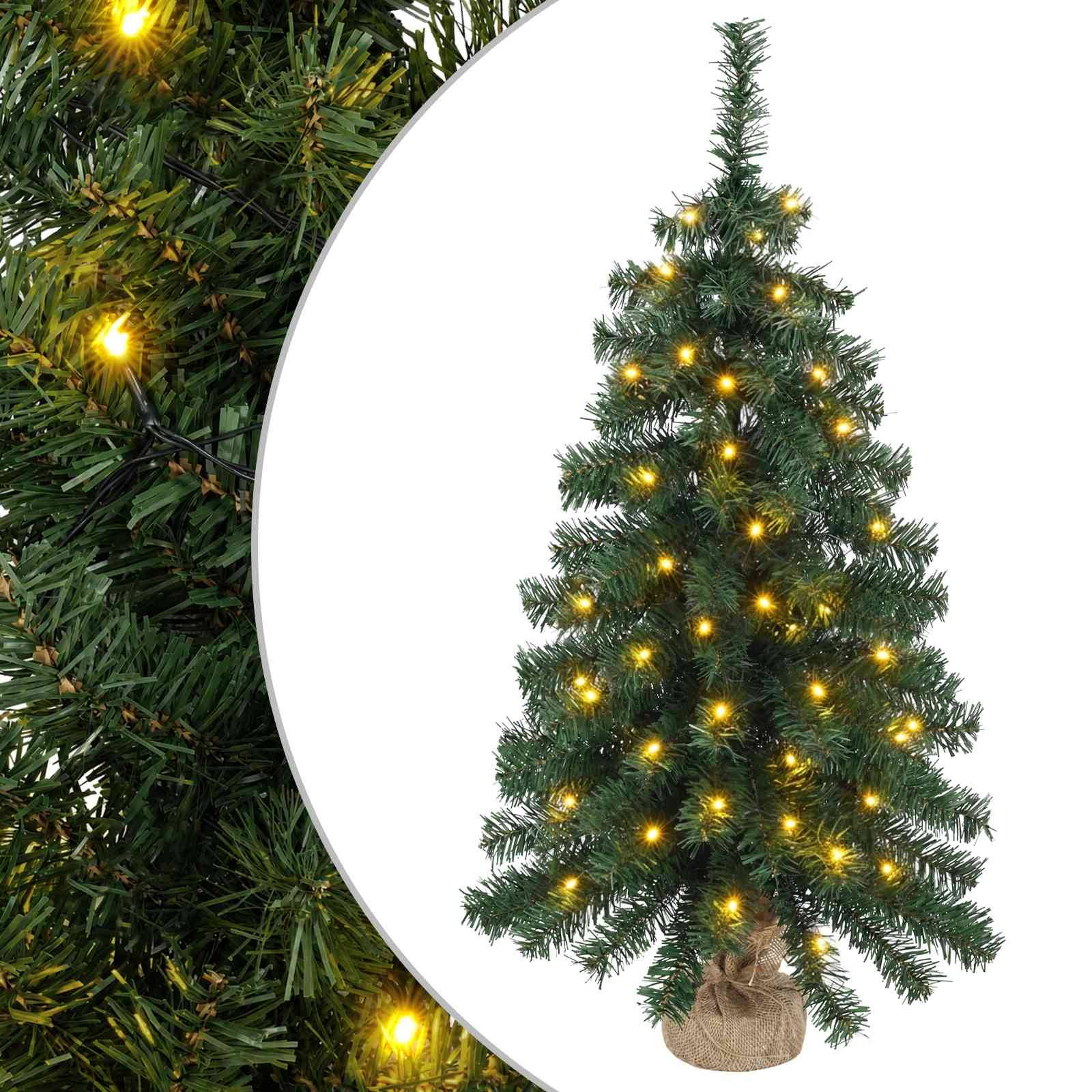 Kleiner künstlicher Weihnachtsbaum mit warmweißen Lichtern und juteumwickeltem Fuß, vor einem detaillierten Immergrün-Hintergrund gezeigt.