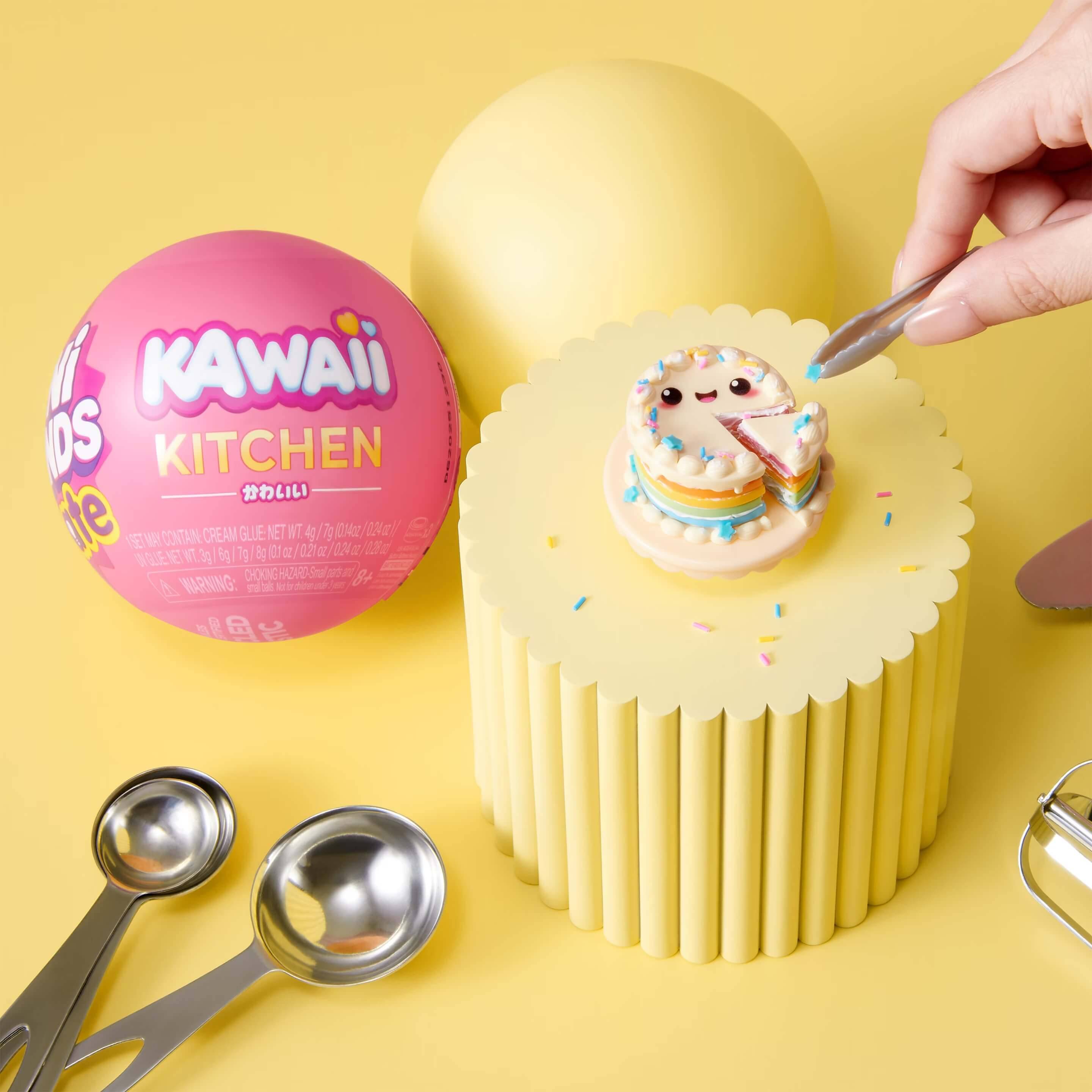 Eine Hand platziert einen Löffel voll neben einem kleinen niedlichen Kuchen auf einem gelben Dessertständer, mit einem rosa „KAWAII KITCHEN" Ball und Messlöffeln in der Nähe.
