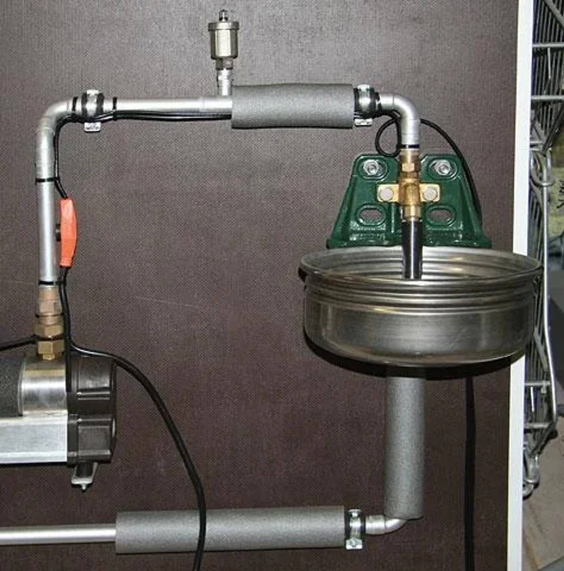 Eine kleine mechanische Pumpenanlage mit Metallrohren, Ventilen und einem rundenSchalenartigen Behälter, der an einer grünen Befestigungshalterung an einer an der Wand montierten Ausstellung angebracht ist.
