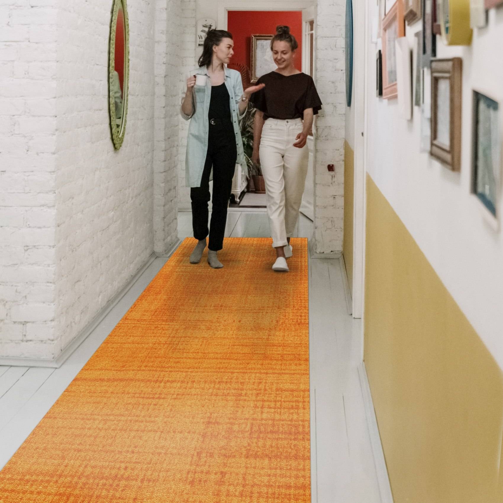 Zwei Frauen gehen und unterhalten sich einen engen Flur hinunter mit einem orangefarbenen Läufer, einer weißen Ziegelmauer und gerahmten Kunstwerken.
