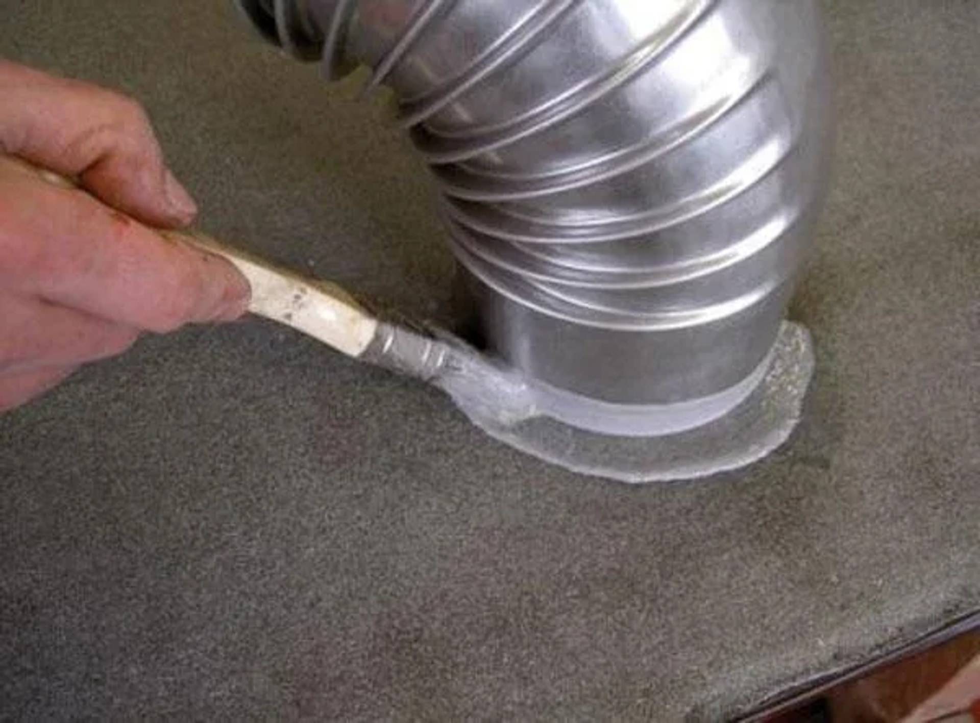 Hand, die Dichtungsmittel um einen flexiblen Metallkanal herum aufträgt, wo dieser auf eine Arbeitsplatte oder Bodenöffnung trifft.