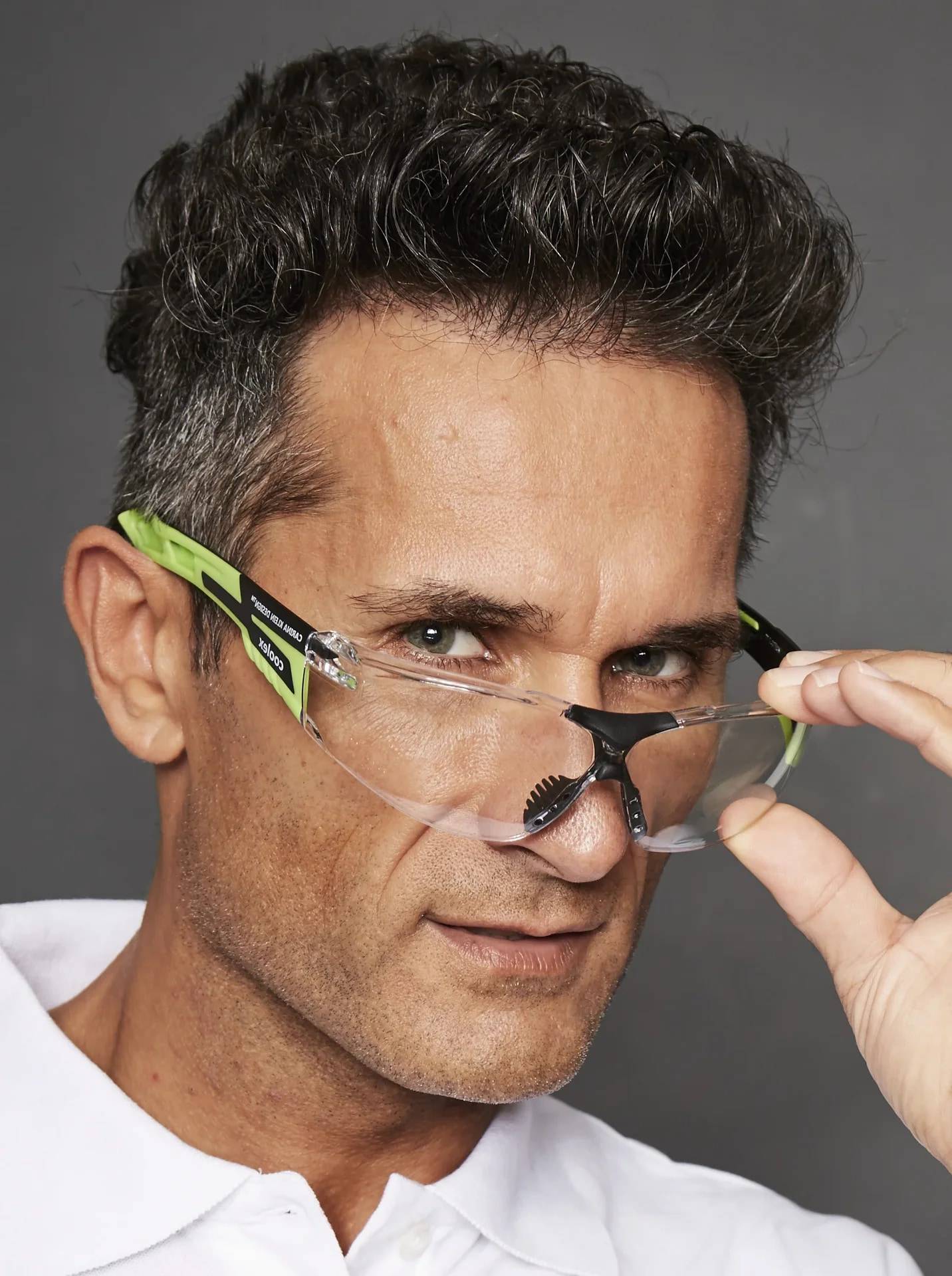 Nahaufnahme eines Mannes in einem weißen Hemd, der eine klare Schutzbrille mit grünen Rahmen vor einem grauen Hintergrund anpasst.