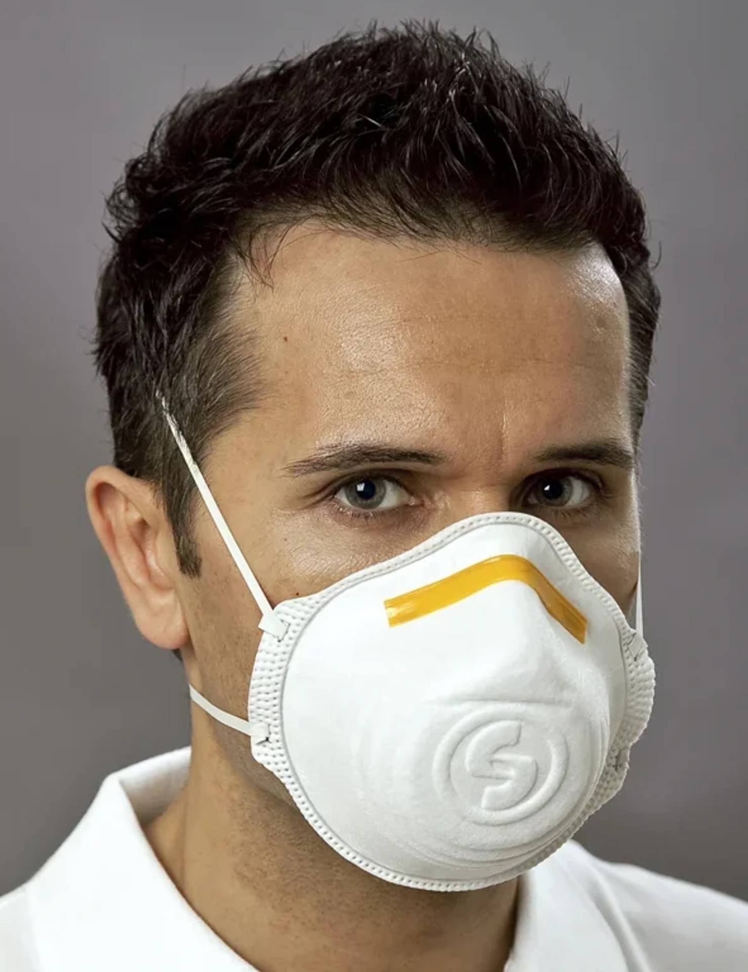 Nahaufnahmeportrait eines Mannes, der eine weiße Schutzgesichtsmaske trägt, in die Kamera schaut vor einem einfarbigen grauen Hintergrund.